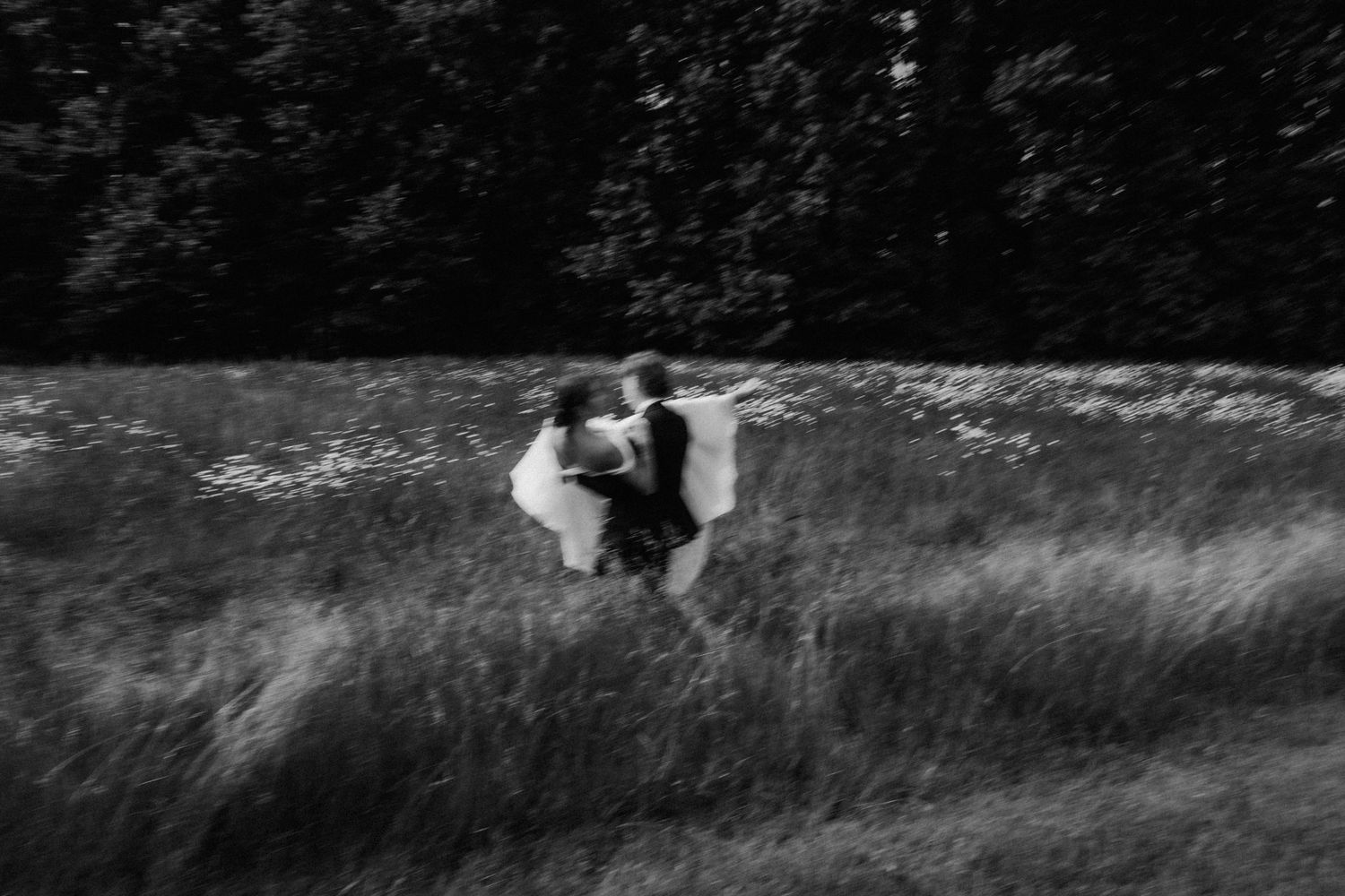 Black and white artistic blurred photo of bride and groom embracing in tall grass field outdoors.