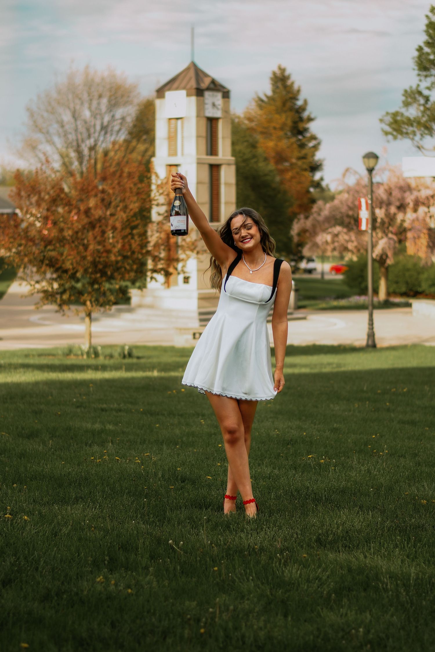 Smiling young woman in white mini dress raising a champagne bottle on a green campus lawn.