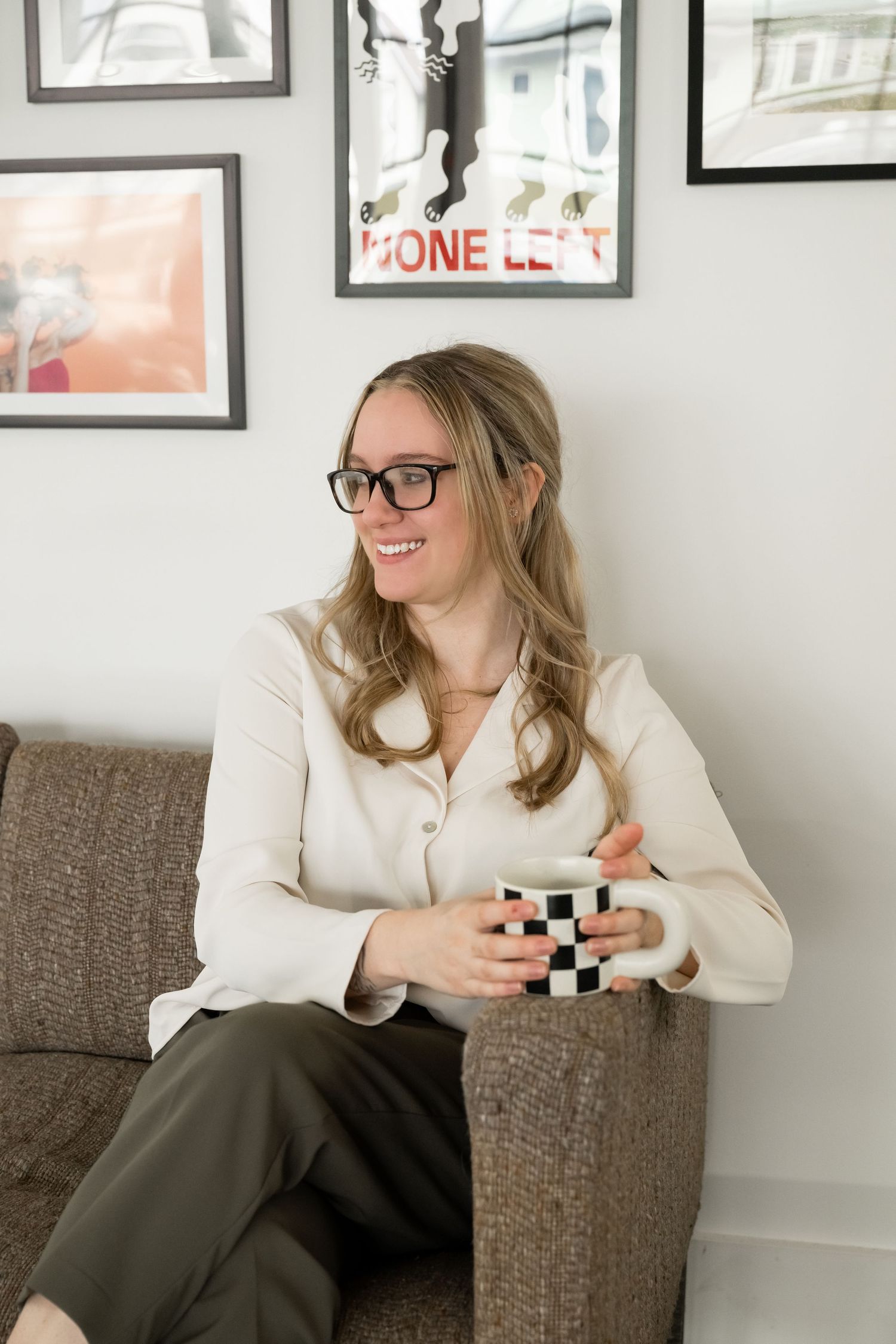 Smiling woman with glasses holding checkered mug sitting on couch with gallery wall behind her.