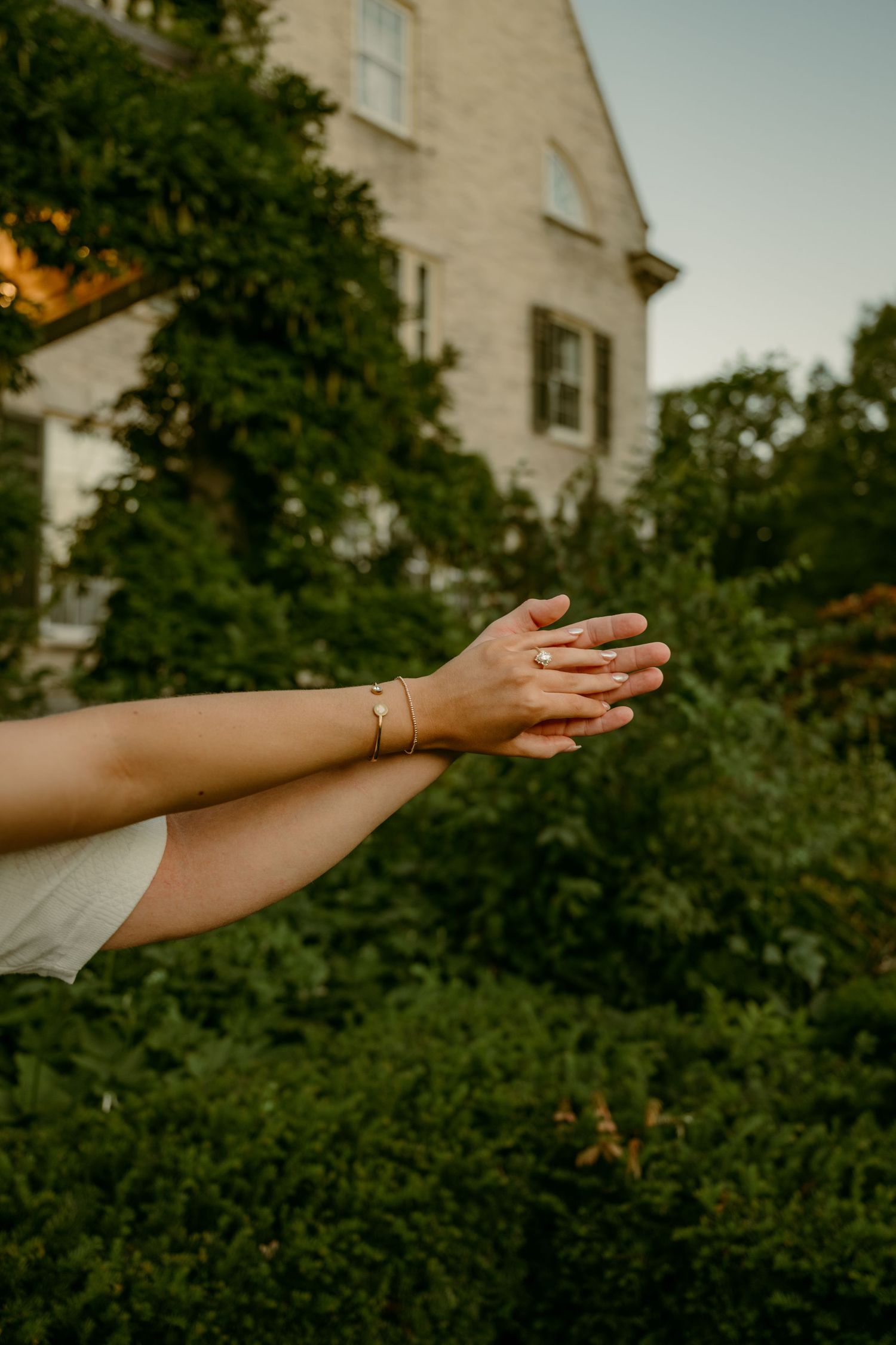 Close-up of a woman's hands with a delicate gold bracelet posed gracefully outdoors near a lush garden.