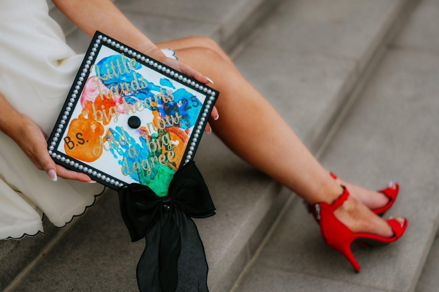Graduate sitting on steps holding colorful decorated mortarboard cap with black bow, wearing red heels.