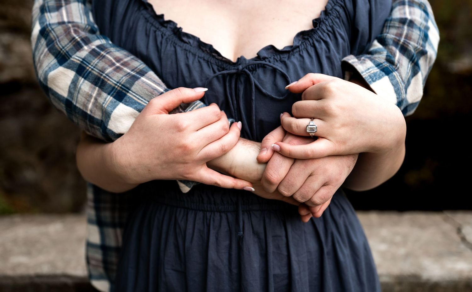 Close-up of two people holding hands, one wearing an engagement ring, in a romantic outdoor setting.