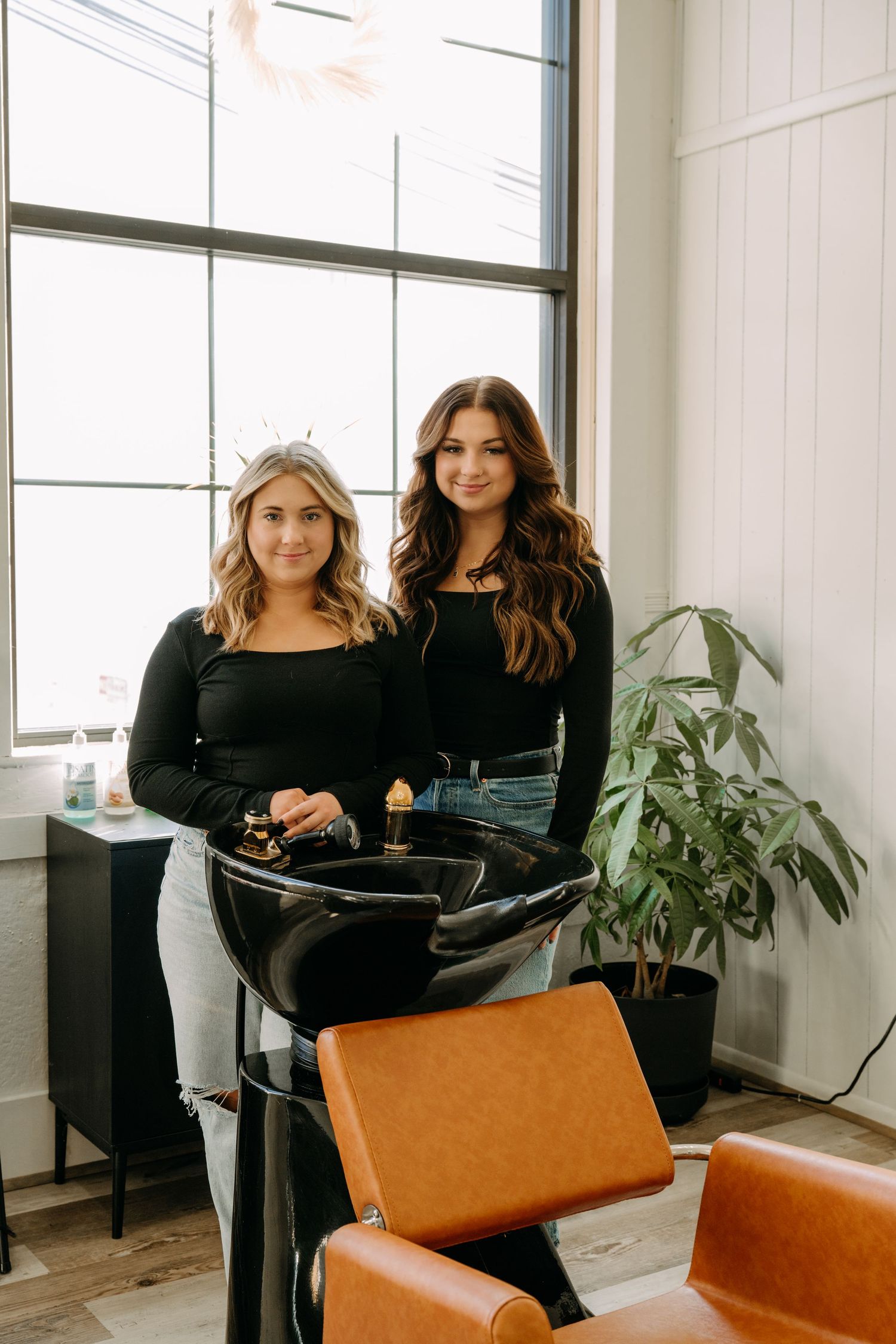 Two female hair stylists in black outfits smiling beside a shampoo bowl in a bright, modern salon interior.