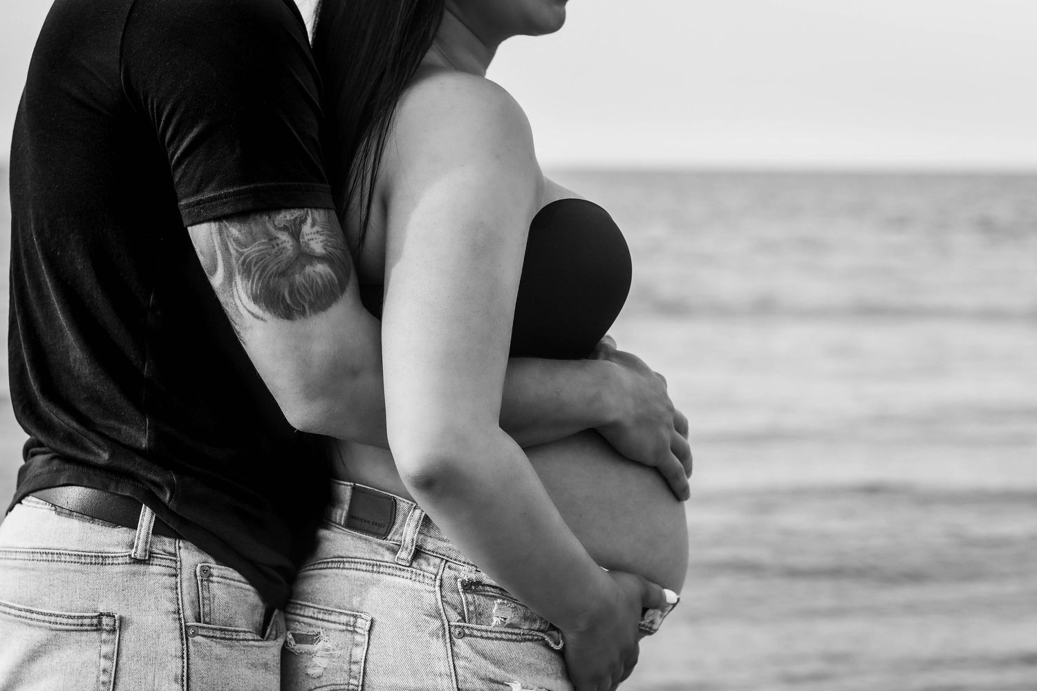 Black and white photo of tattooed couple embracing on beach, man holding pregnant woman's belly by ocean.