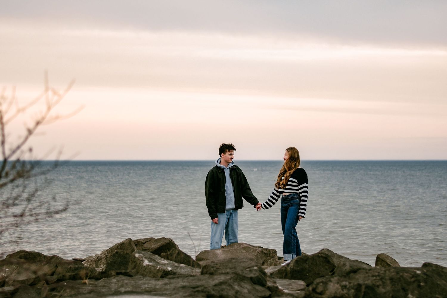 Young couple holding hands on rocky shoreline at sunset with calm ocean behind them.