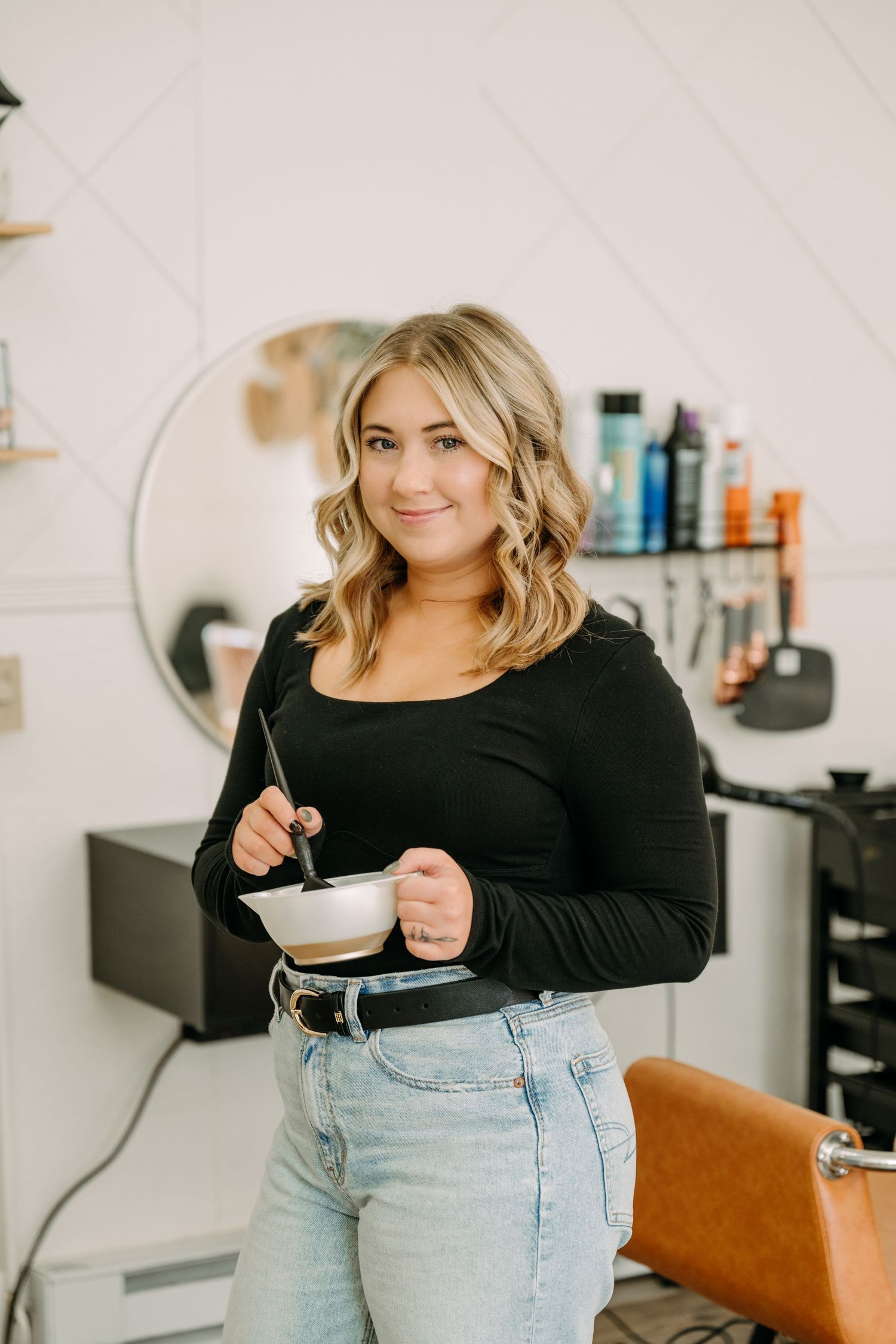 Blonde hairstylist in black top holding color bowl and brush in salon.