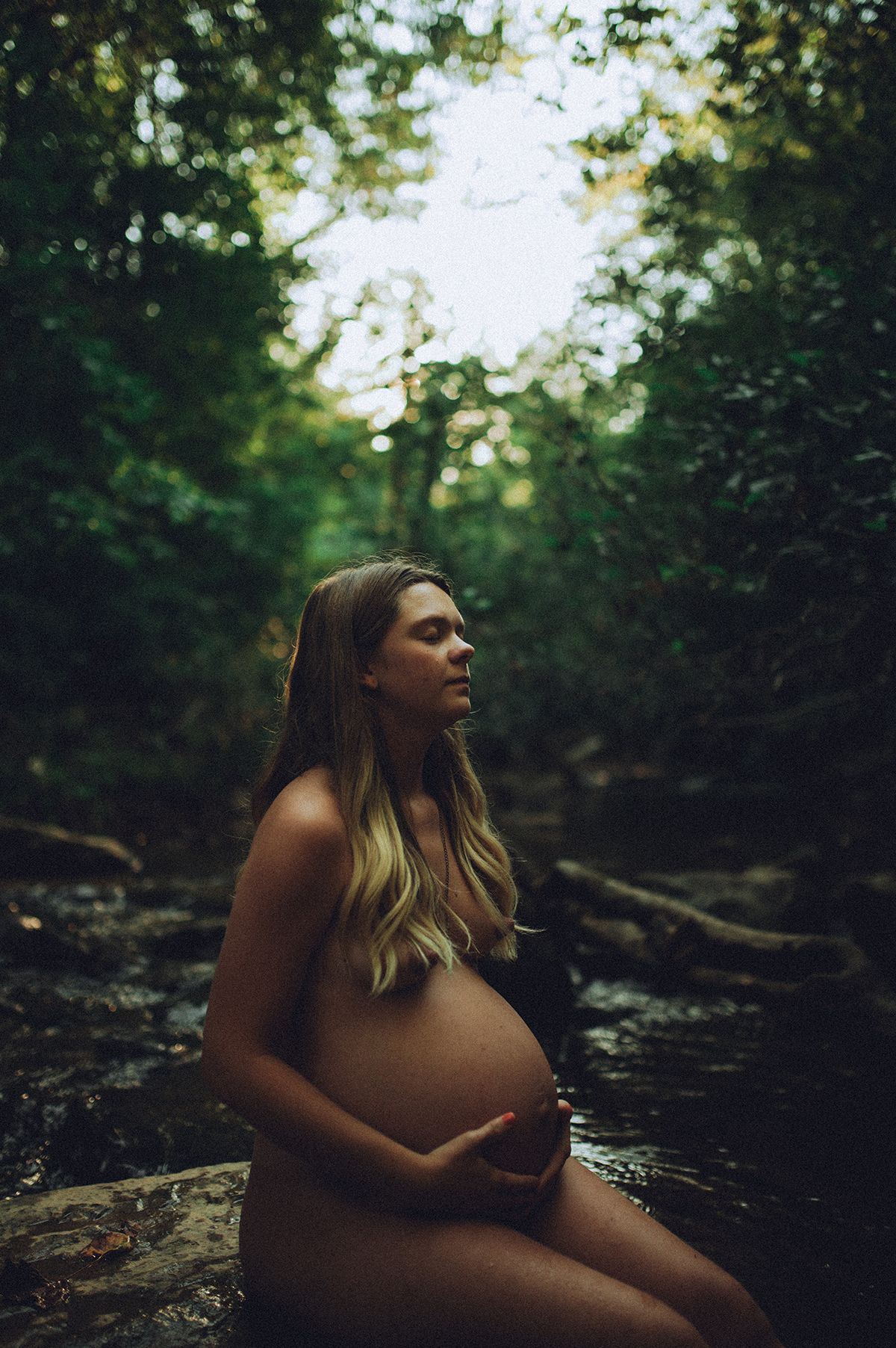 Moody forest maternity boudoir portraits showing a woman reclining on rocks in dappled natural lighting near Spartanburg, SC.