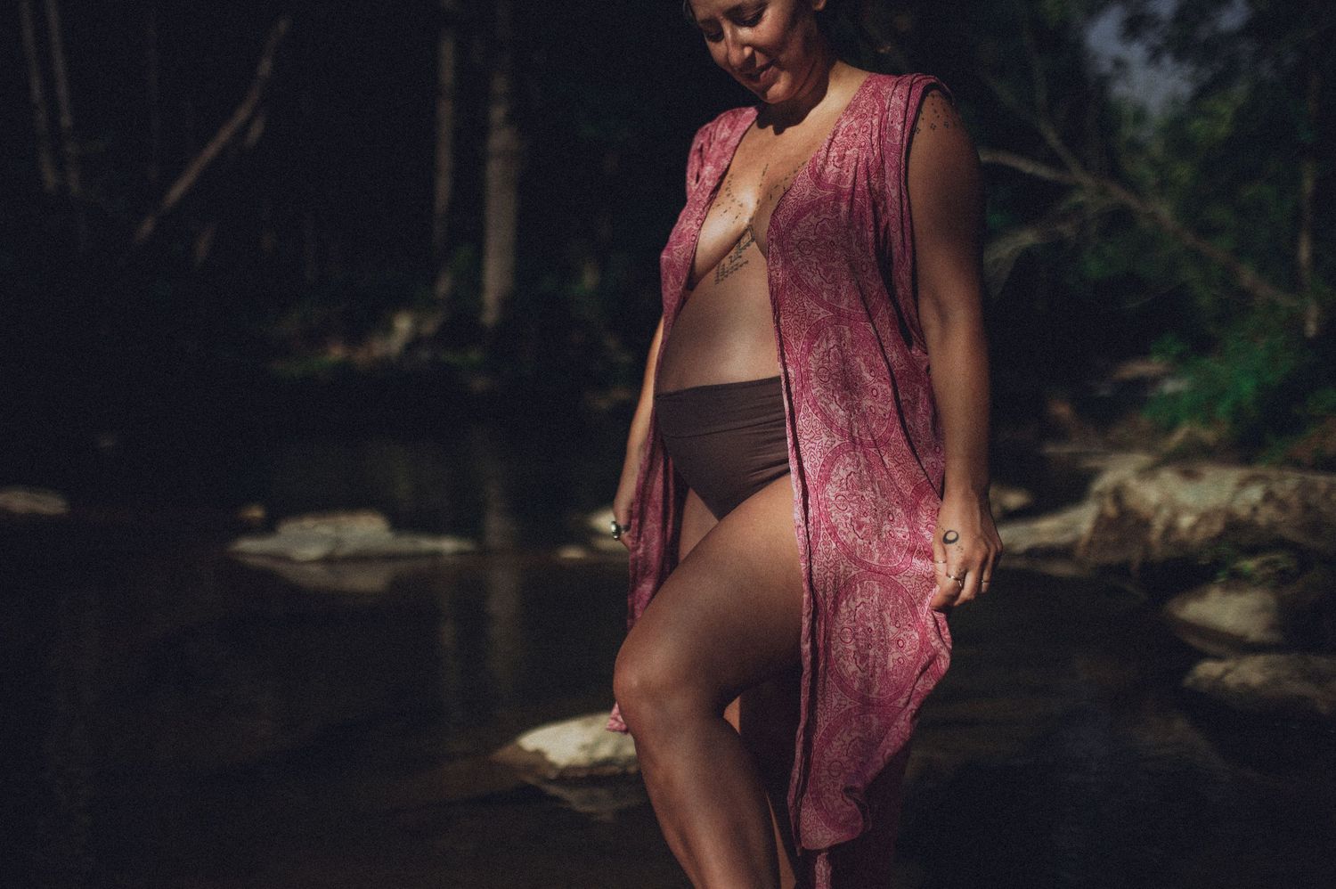 A woman wearing a pink flowy dress poses in a forest setting with dappled sunlight filtering through the trees in NC. Maternity portrait.