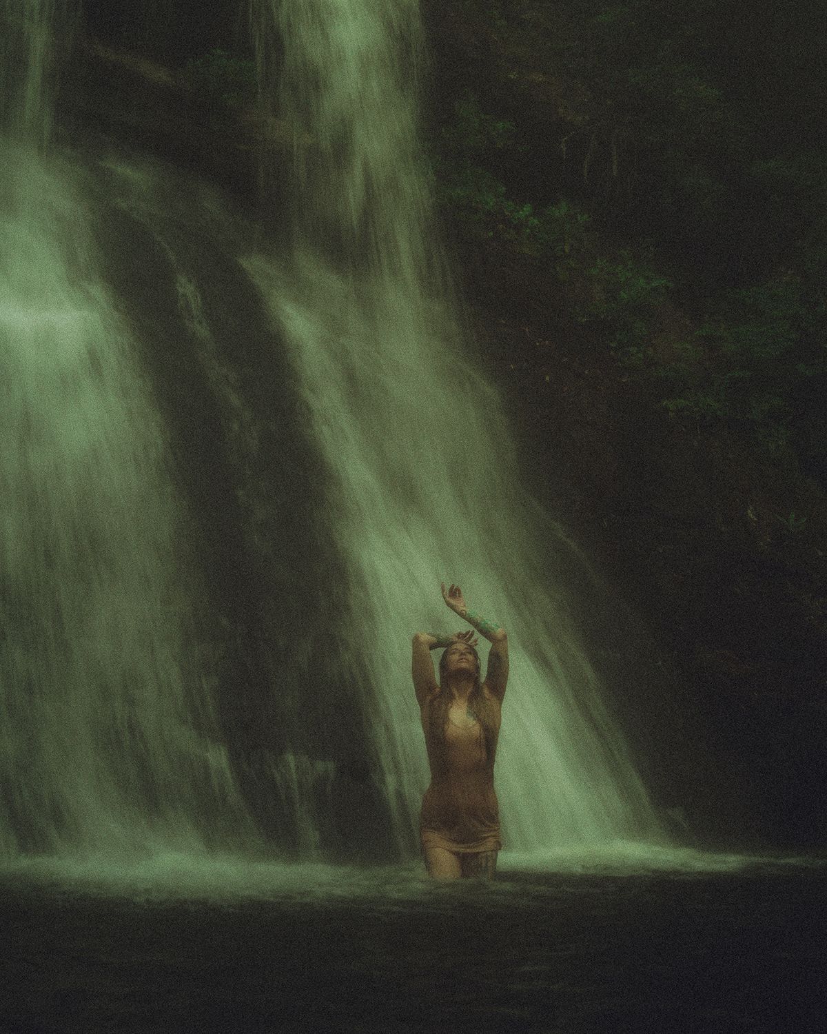 A figure stands silhouetted against a cascading waterfall in a dark, moody forest setting creating a mystical boudoir portrait in NC.