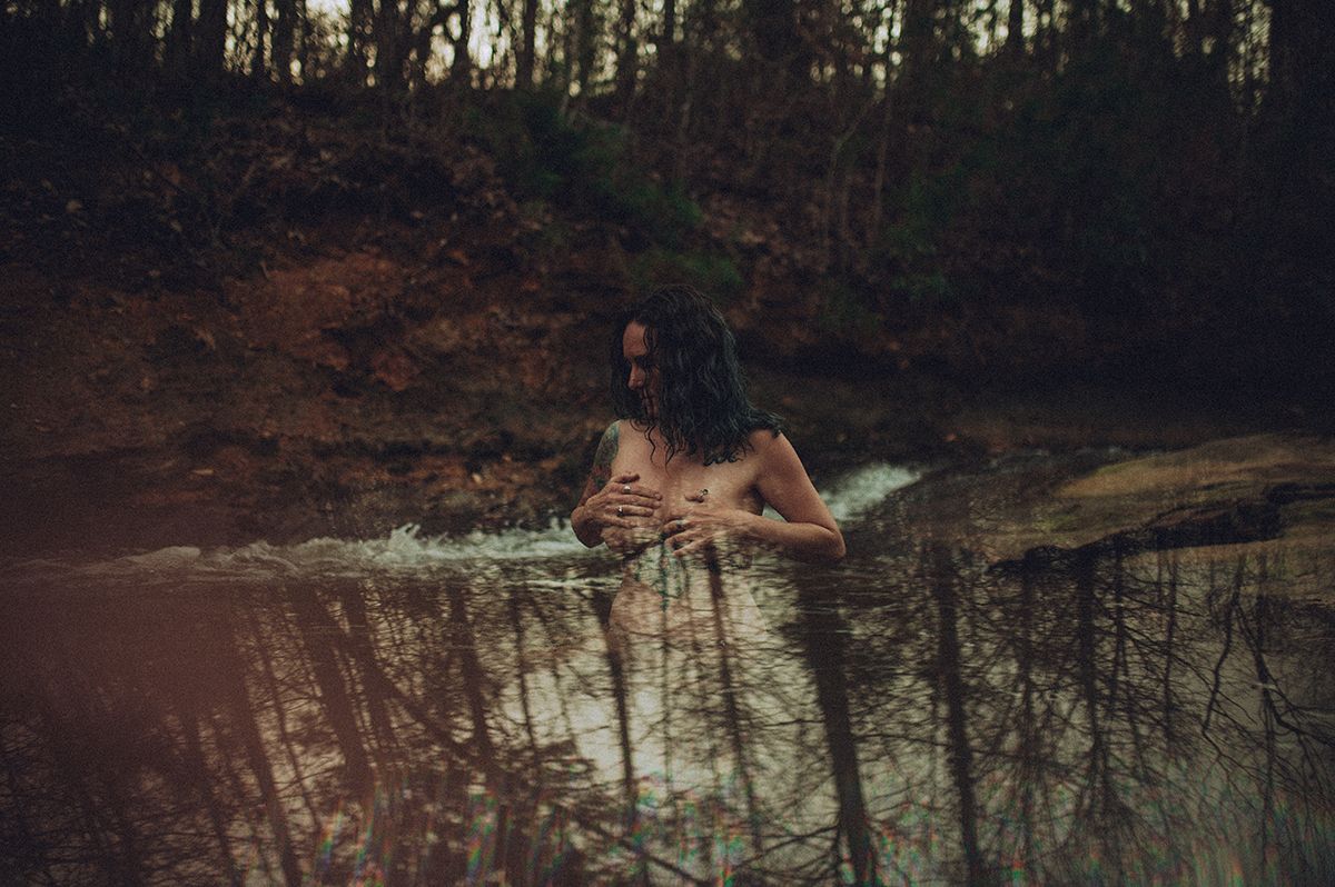 A person swims in a tranquil forest pond surrounded by trees during sunset creating a serene natural boudoir scene in Spartanburg, SC.