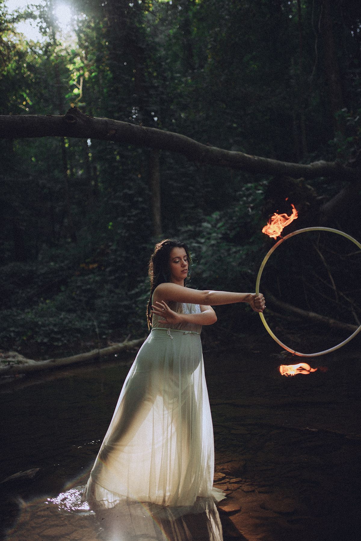 A figure in a flowing white dress performs fire dancing in a dark wooded setting with a flaming hoop in NC river. Maternity Portrait.