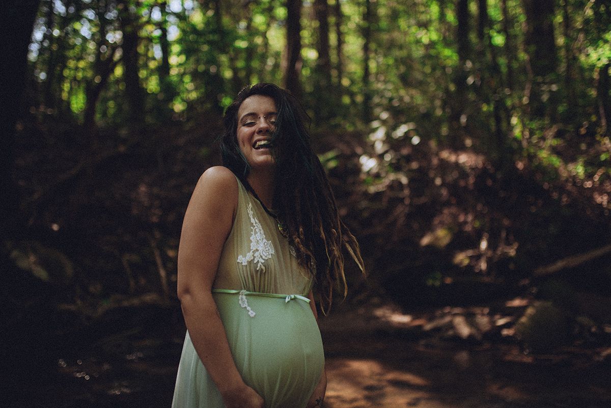 Maternity boudoir portrait in a sage green dress posing in a sunlit forest setting with dappled light filtering through the trees in NC.