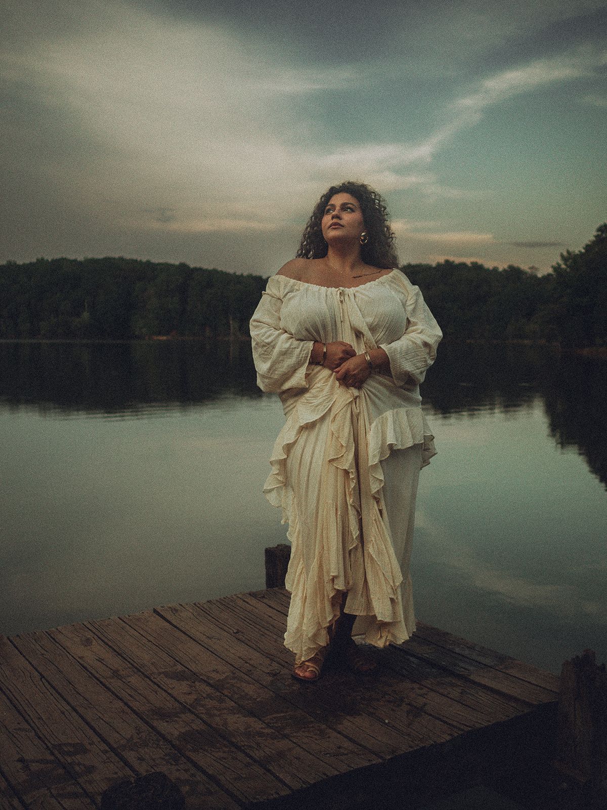 A figure in a vintage white dress poses dramatically on a wooden dock by a misty lake at dusk in Spartanburg, SC.