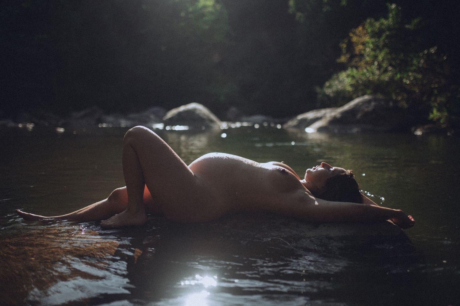 Artistic nude maternity boudoir portrait of a woman reclining on rocks in a forest stream with sunlight filtering through the trees in NC.
