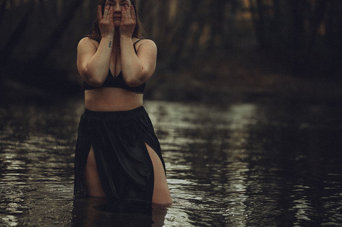 Silhouetted figure in dark dress stands in still water during moody forest photoshoot in North Carolina.