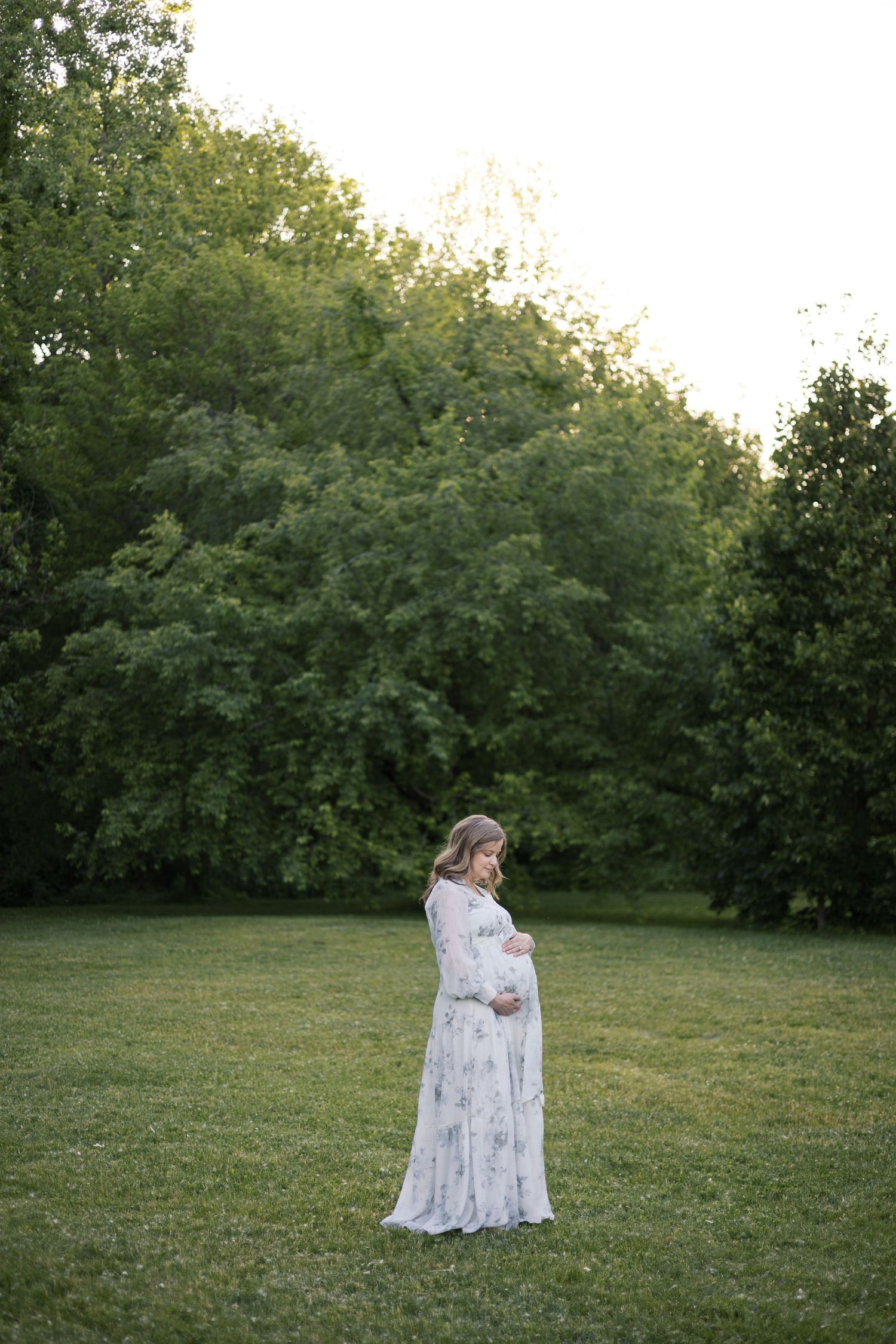 Pregnant woman in flowing floral dress cradling baby bump while standing alone in serene green park by Katy Rose Photography.