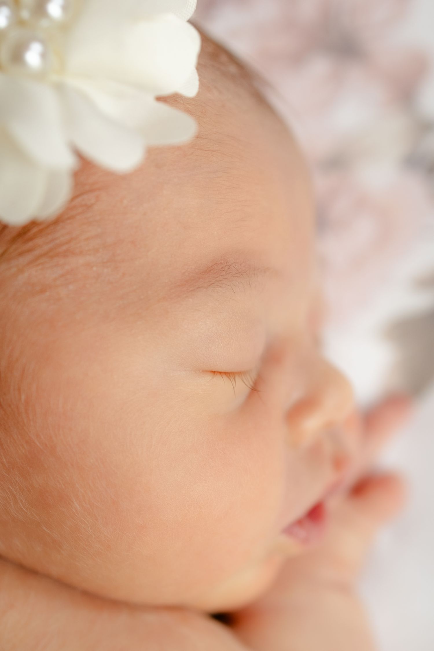 Peaceful sleeping newborn baby girl wearing a white flower headband with pearl accents in a soft portrait photo during newborn photos