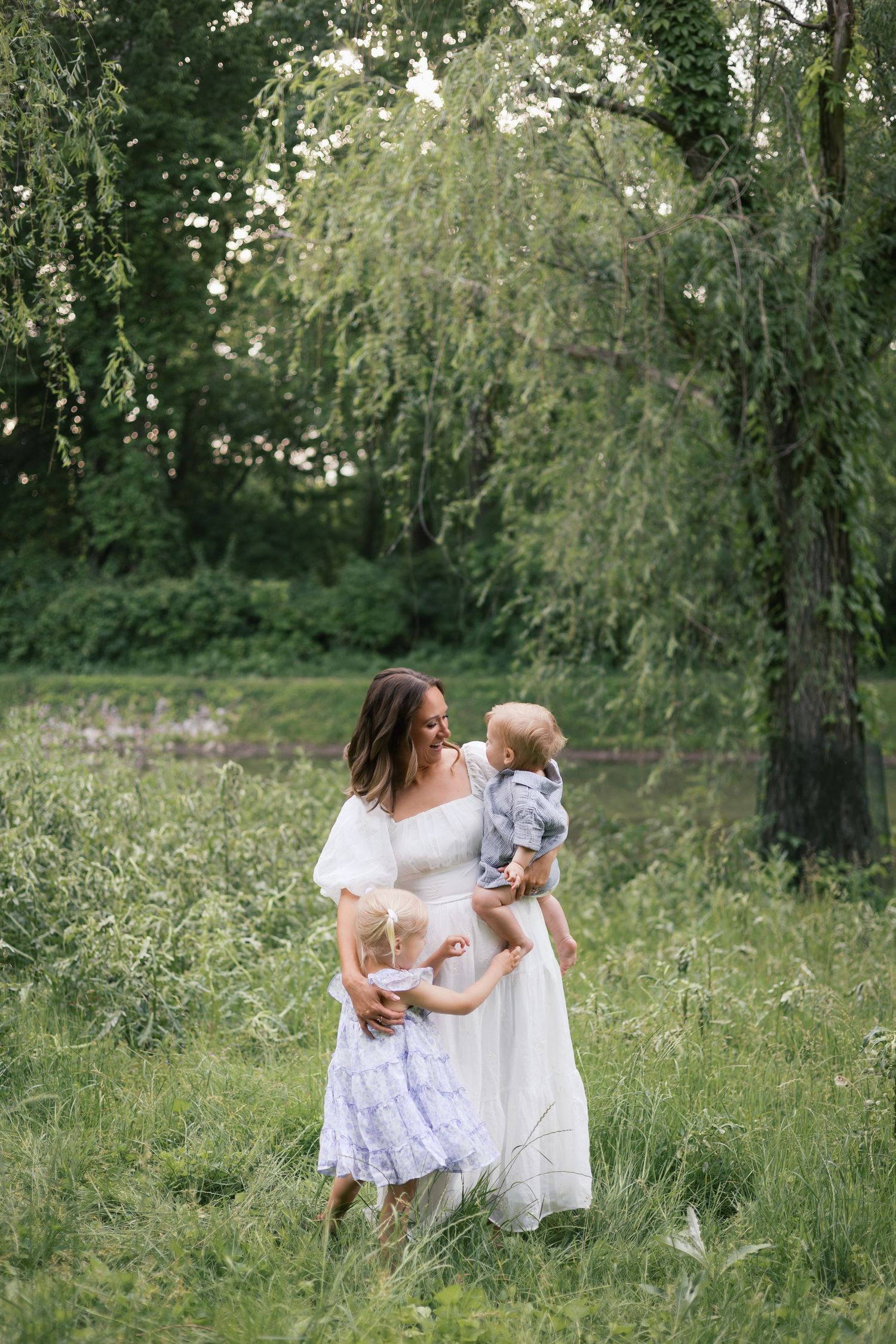 A mother in a white dress holds her baby while her young daughter stands beside her in a lush green park.