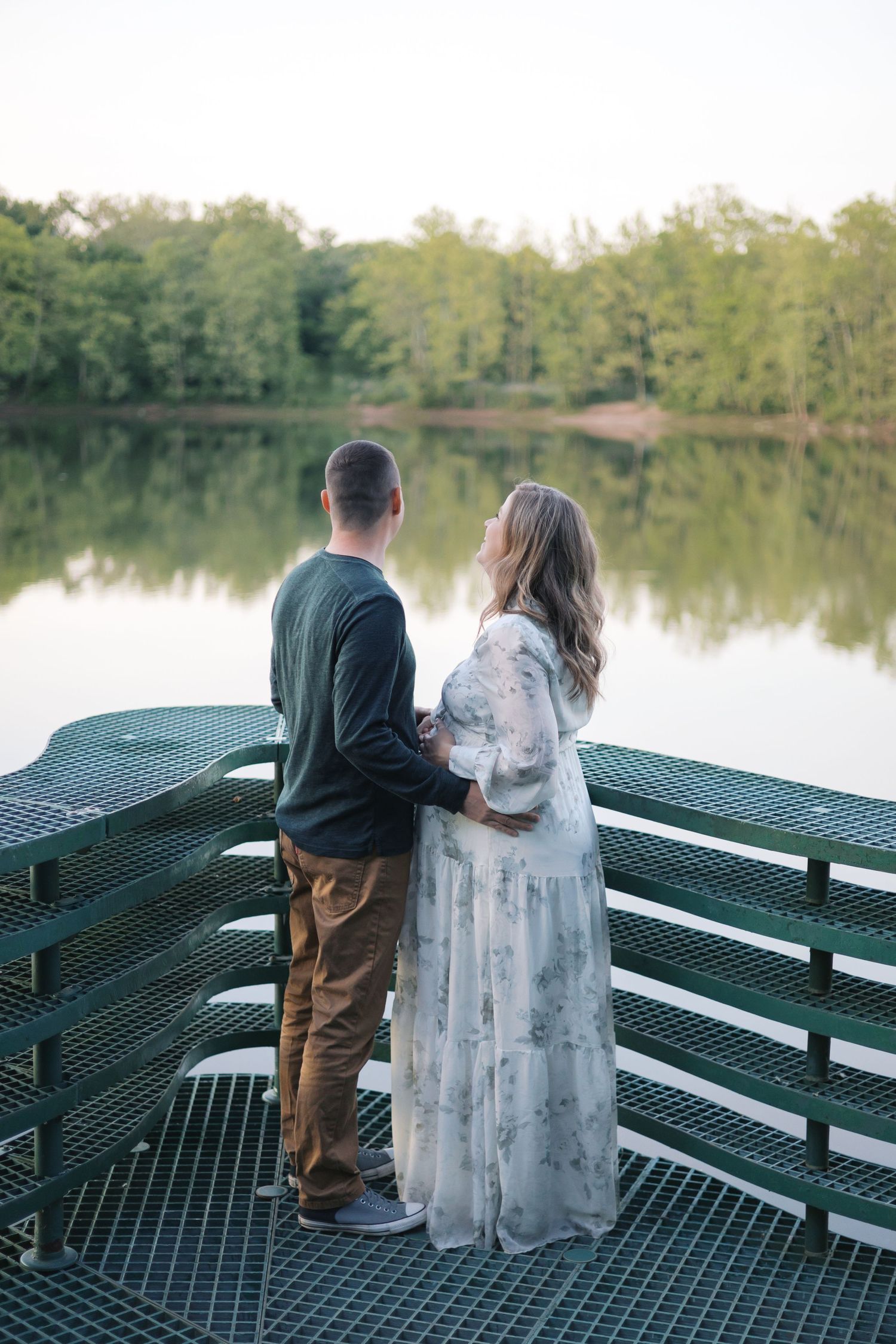 Couple standing on a green metal dock overlooking a calm lake surrounded by lush green trees.