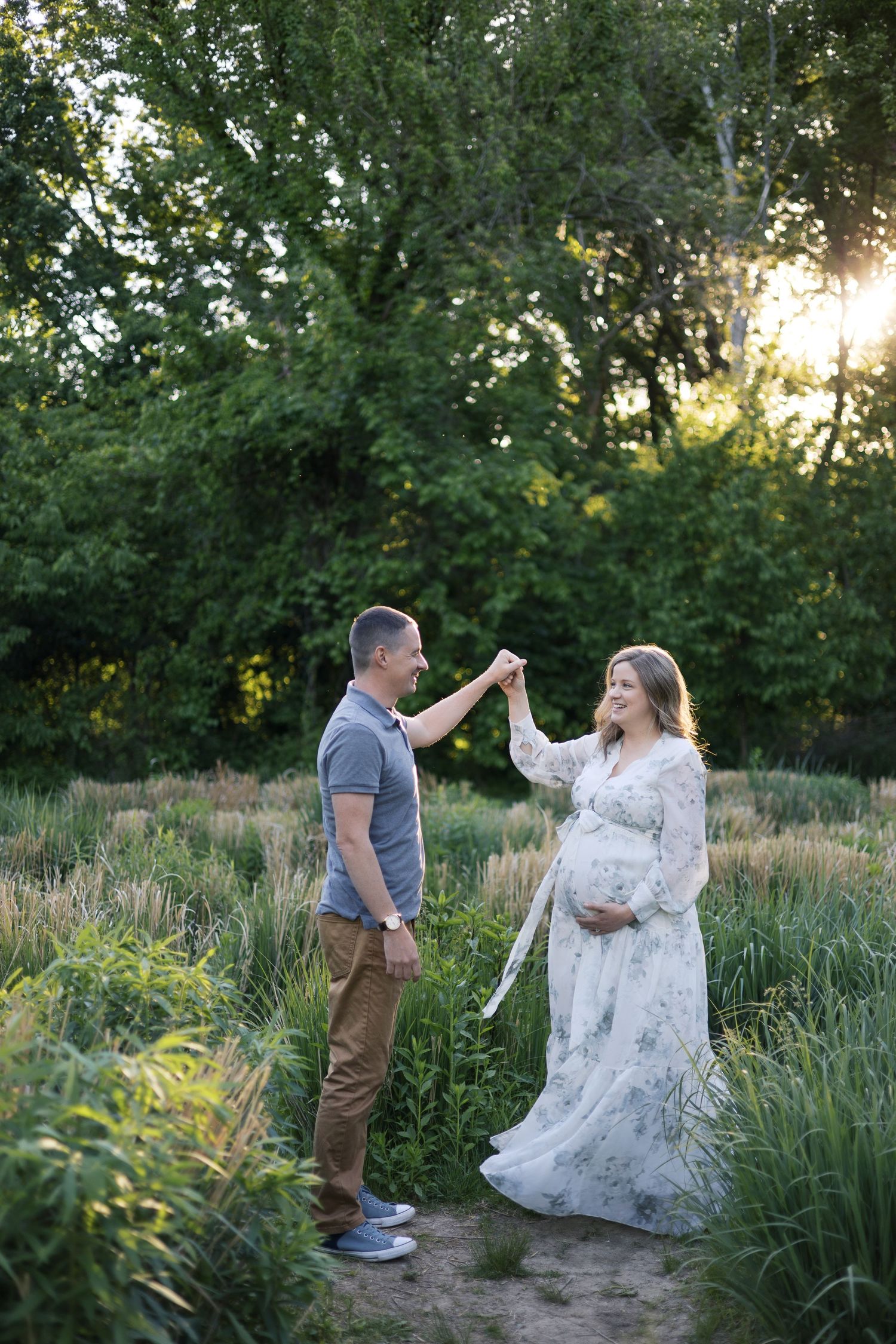 Couple dancing outdoors at sunset, pregnant woman in floral dress holding hands with man in green park setting.