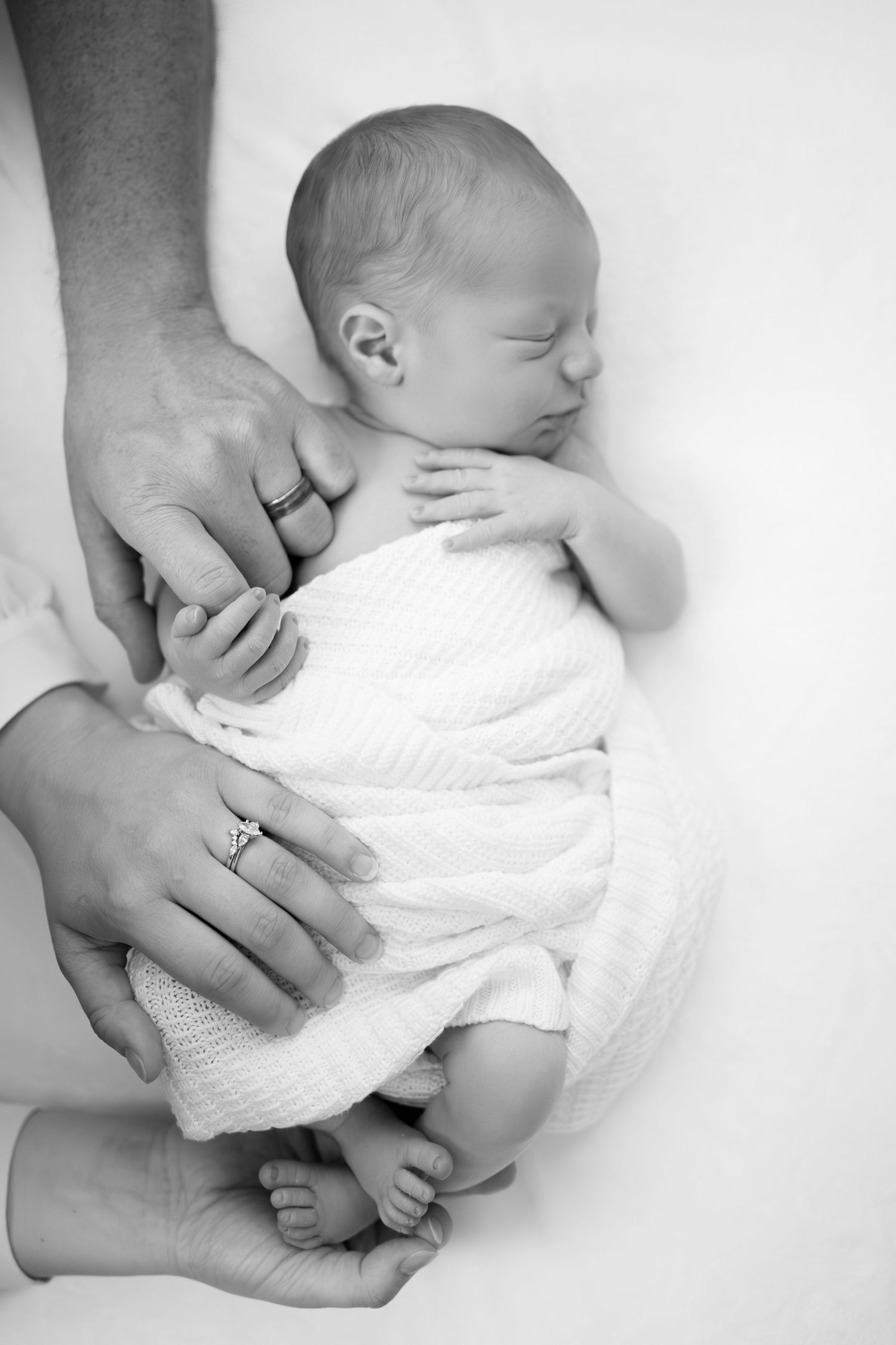 Black and white photo of a sleeping newborn baby wrapped in white cloth, cradled gently by parents hands.