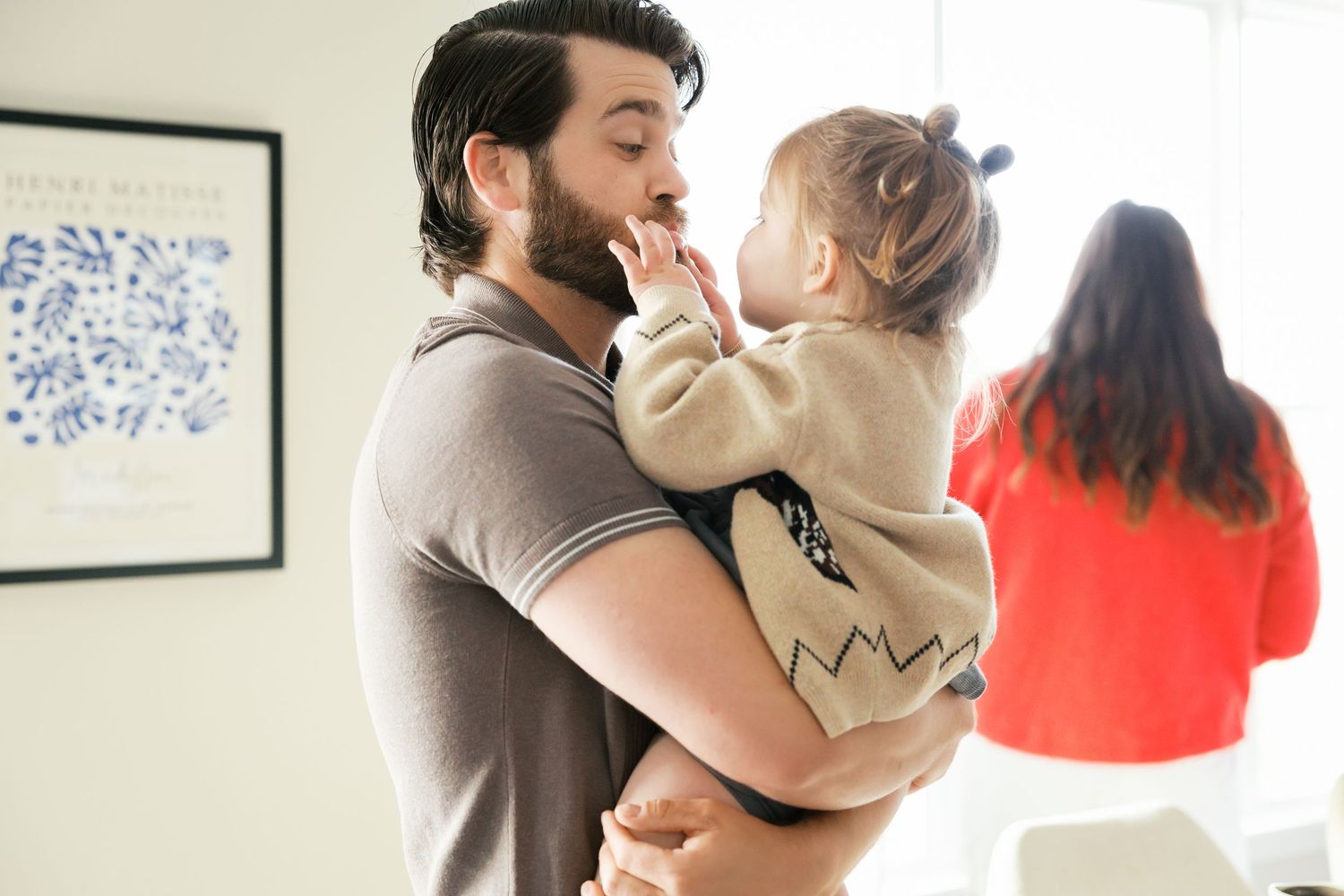 A bearded father holds and smiles at his toddler girl indoors while a woman in red stands in the background.