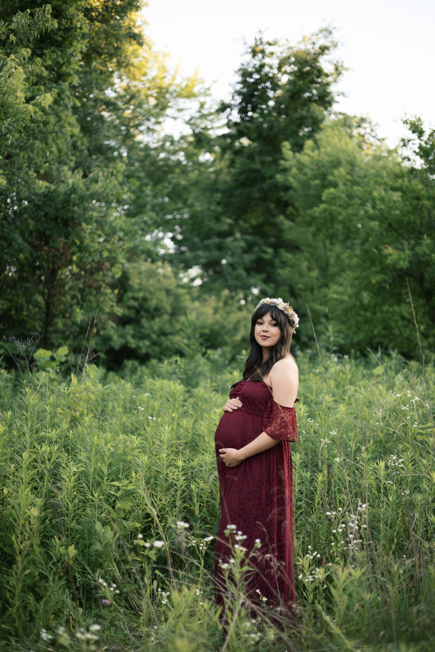 Pregnant woman in burgundy lace gown and floral crown standing in lush green field for maternity photo session in Danville, Indiana.