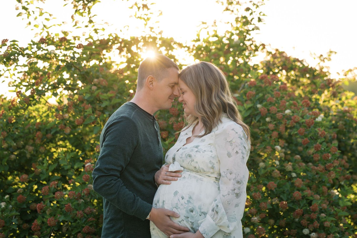 Couple sharing a tender moment outdoors, pregnant woman in floral dress, golden sunset light through blooming bushes.