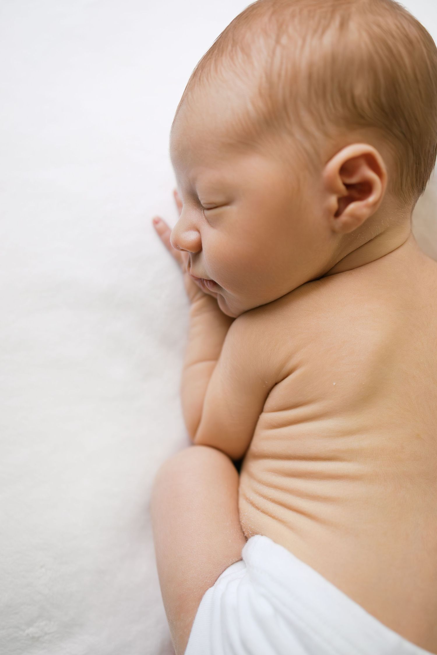A peaceful newborn baby sleeping on a soft white blanket, wearing a diaper, with eyes closed and hand near face.