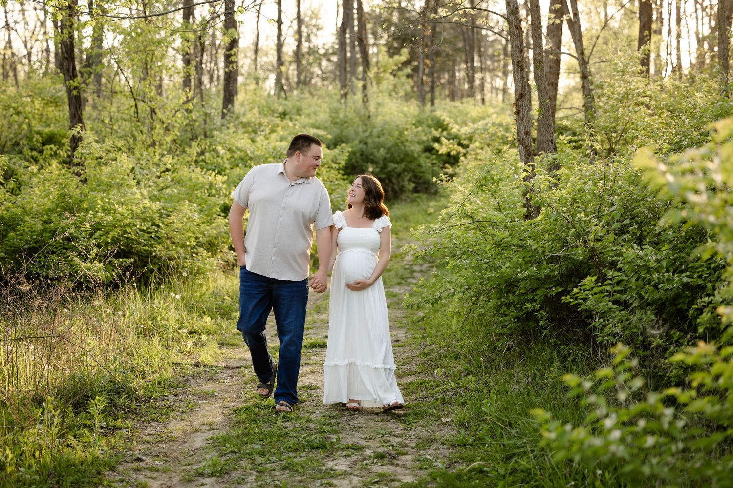 Pregnant couple walking hand in hand on a forest path surrounded by lush green trees during a maternity photo session in Hendricks County.