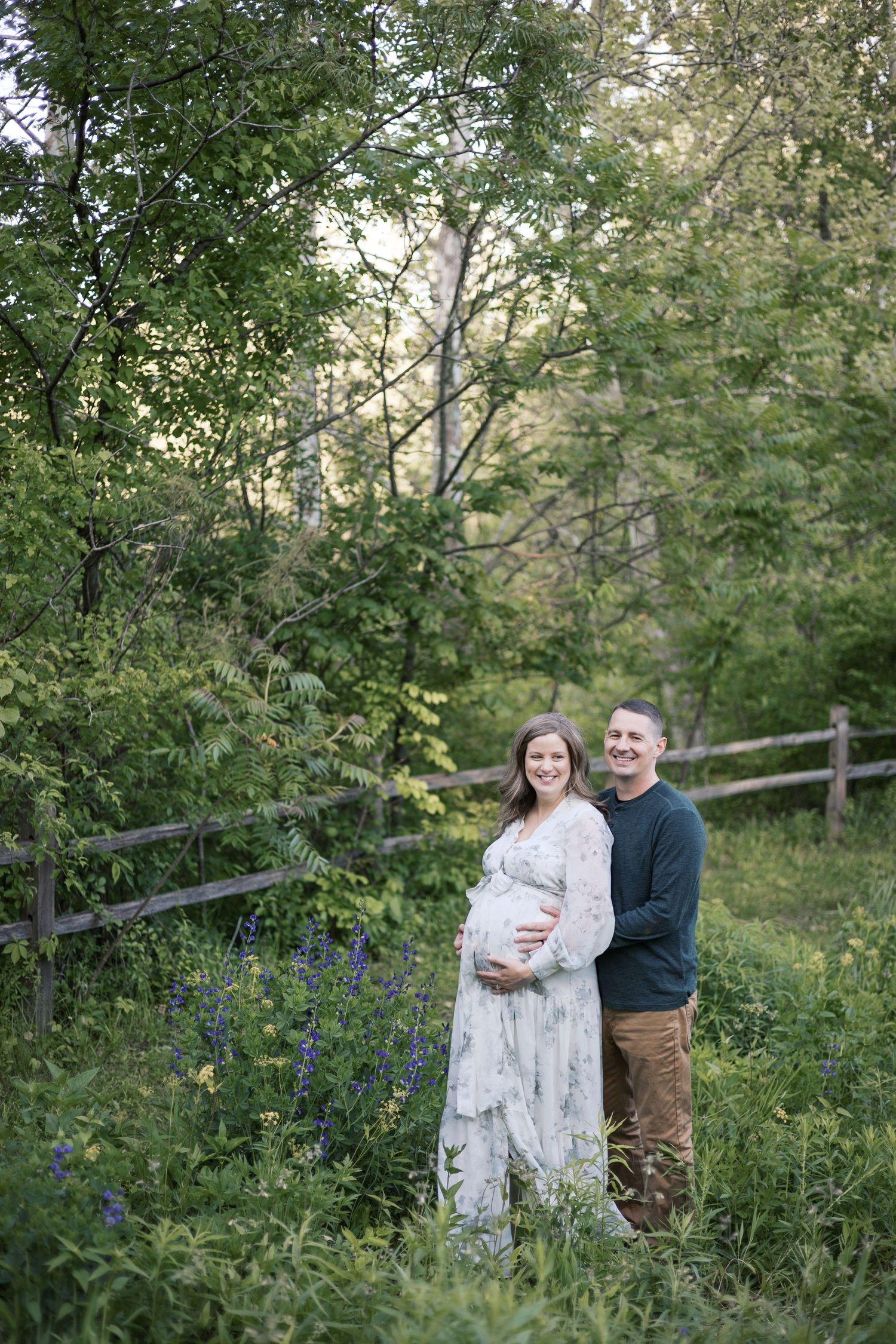 Expecting couple smiling together in a garden, woman in floral dress cradling baby bump for maternity portraits by Katy Rose Photography.