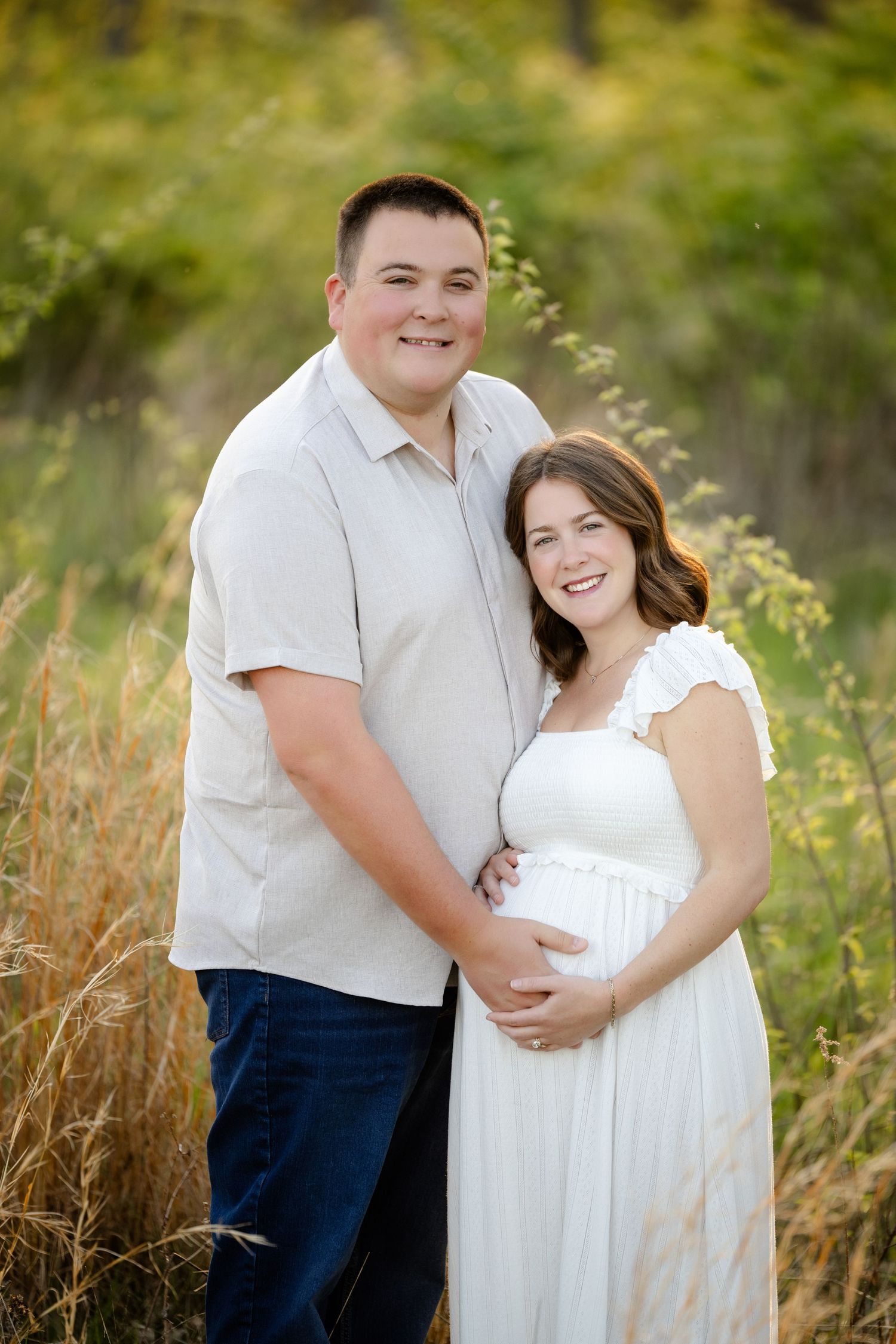 Smiling couple poses in golden field, pregnant woman in white dress cradling baby bump with partner embracing her during maternity session