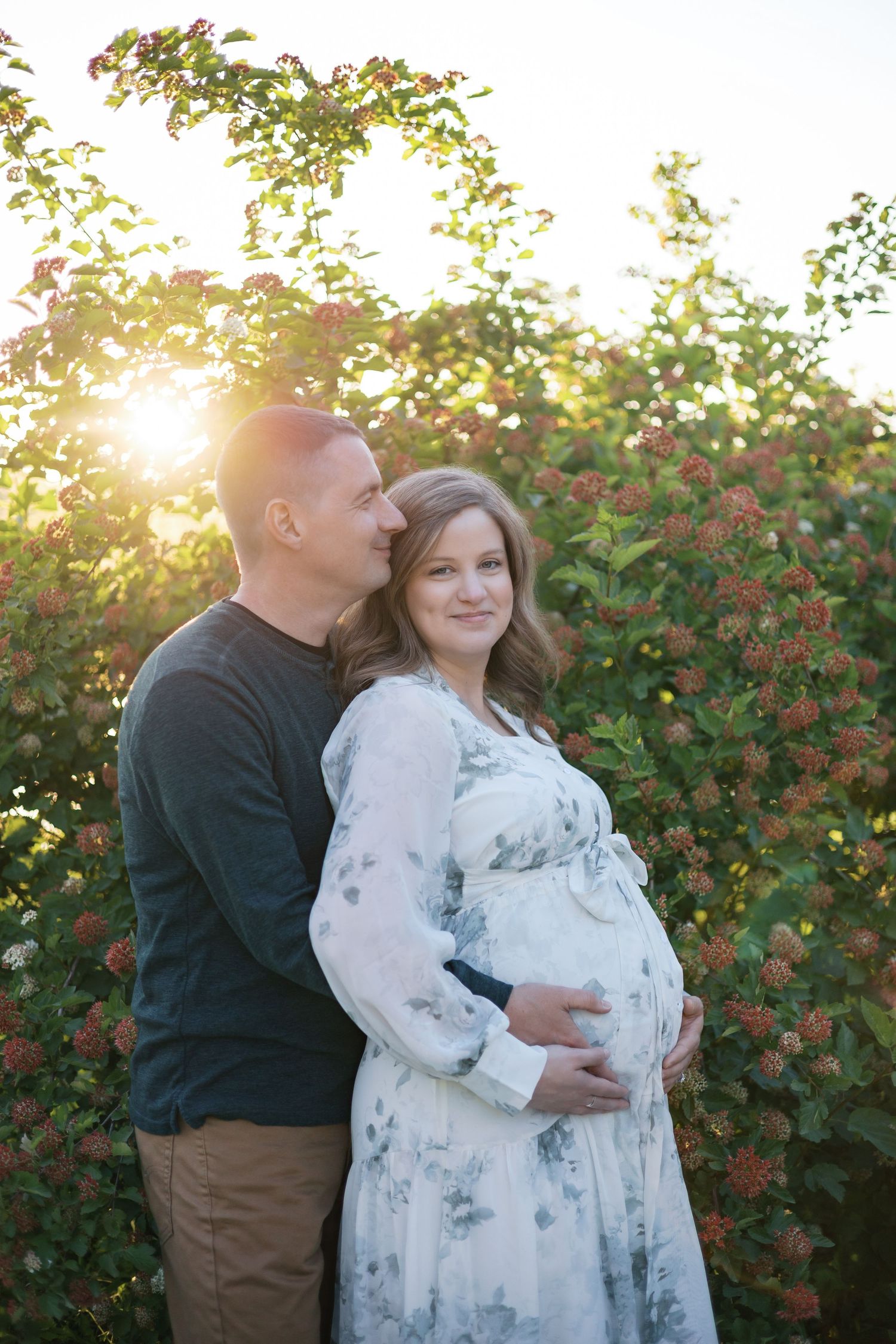Couple embracing in front of blooming flowers with golden sunset light shining behind them during maternity photos by Katy Rose Photography.