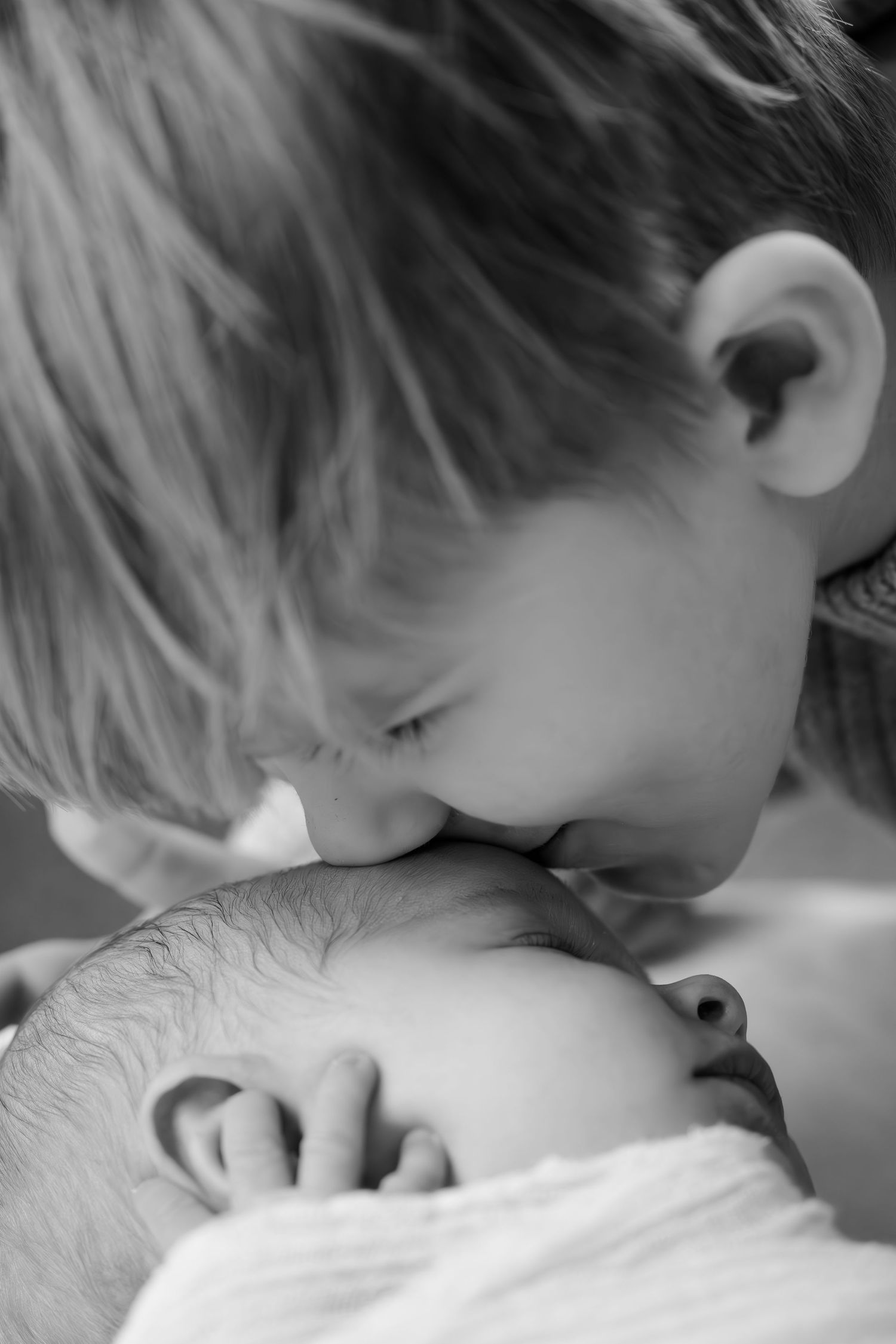 Black and white photo of a young child gently kissing a sleeping newborn baby on the head at Indianapolis in-home newborn session.