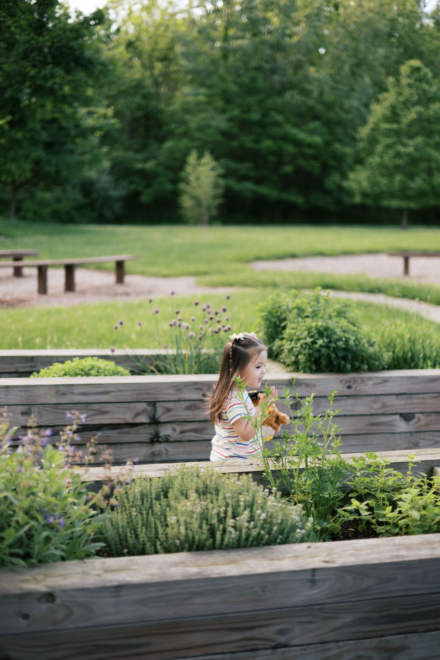 A young girl explores wooden raised garden beds filled with herbs and flowers in a lush green park setting.
