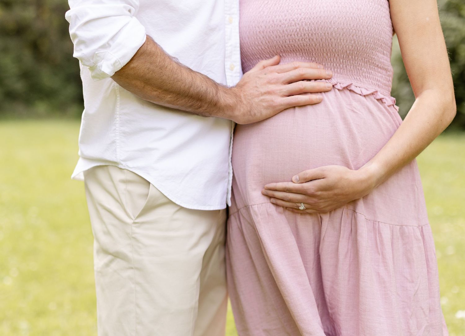 Close-up of couple's hands cradling baby bump in pink maternity dress.