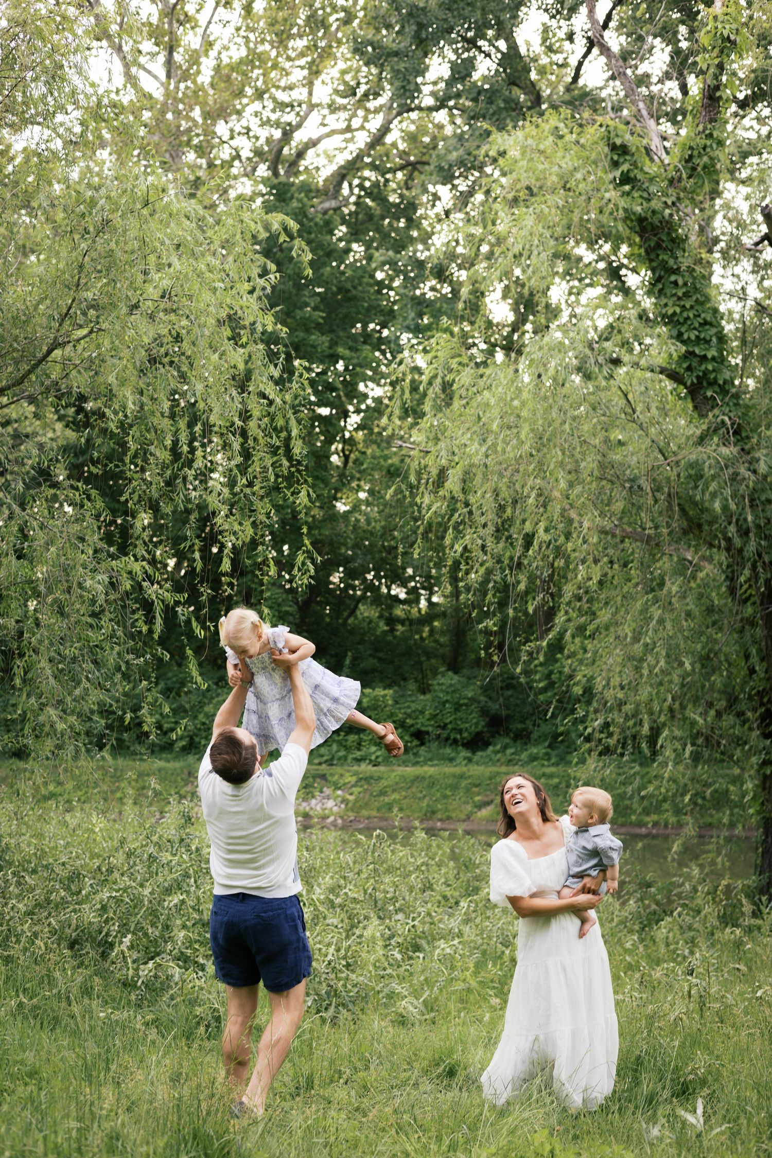 A joyful family plays outdoors in a lush green park, with father lifting a child while mother holds a toddler.