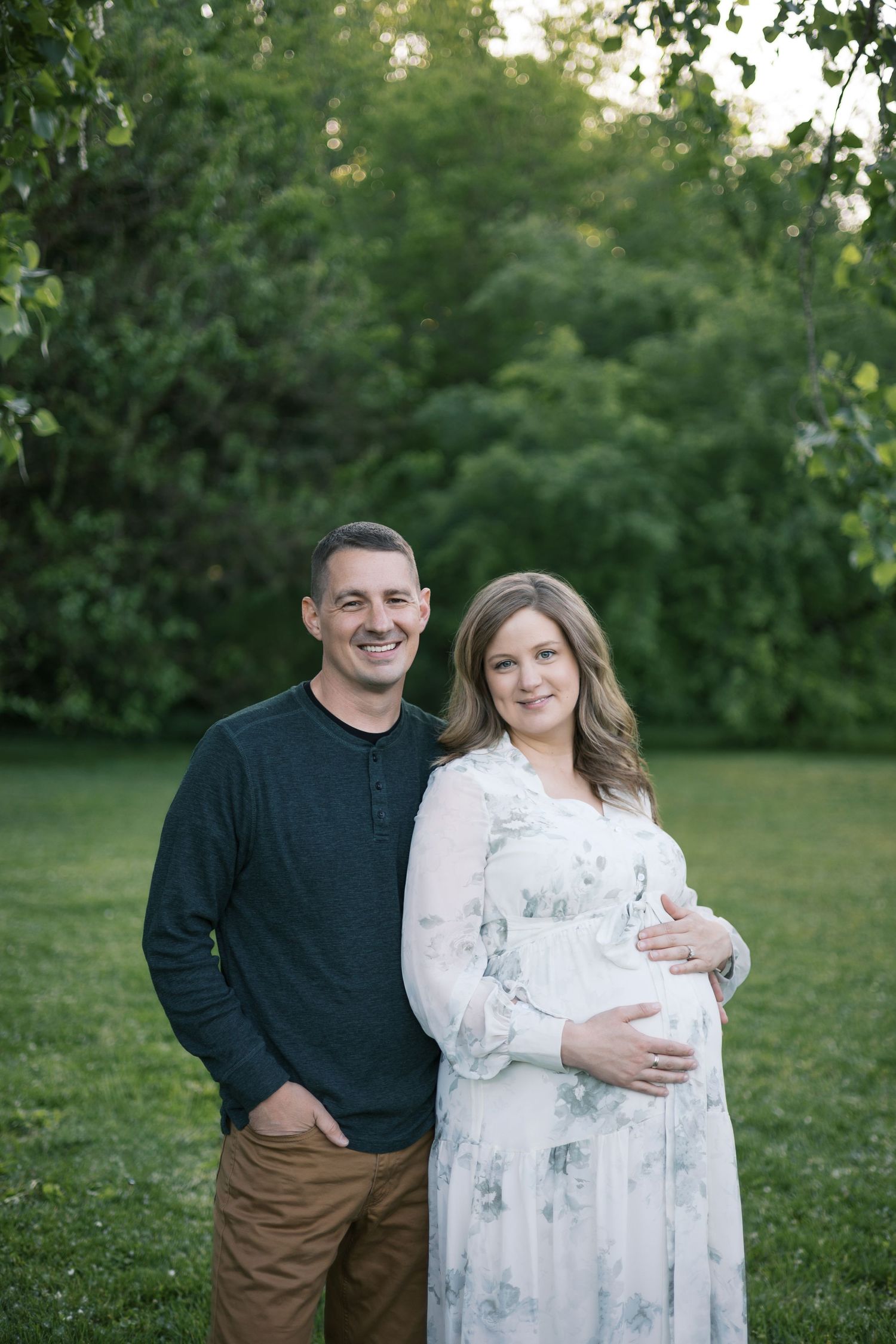 Smiling couple posing together outdoors on green lawn, pregnant woman in floral maxi dress at dusk.