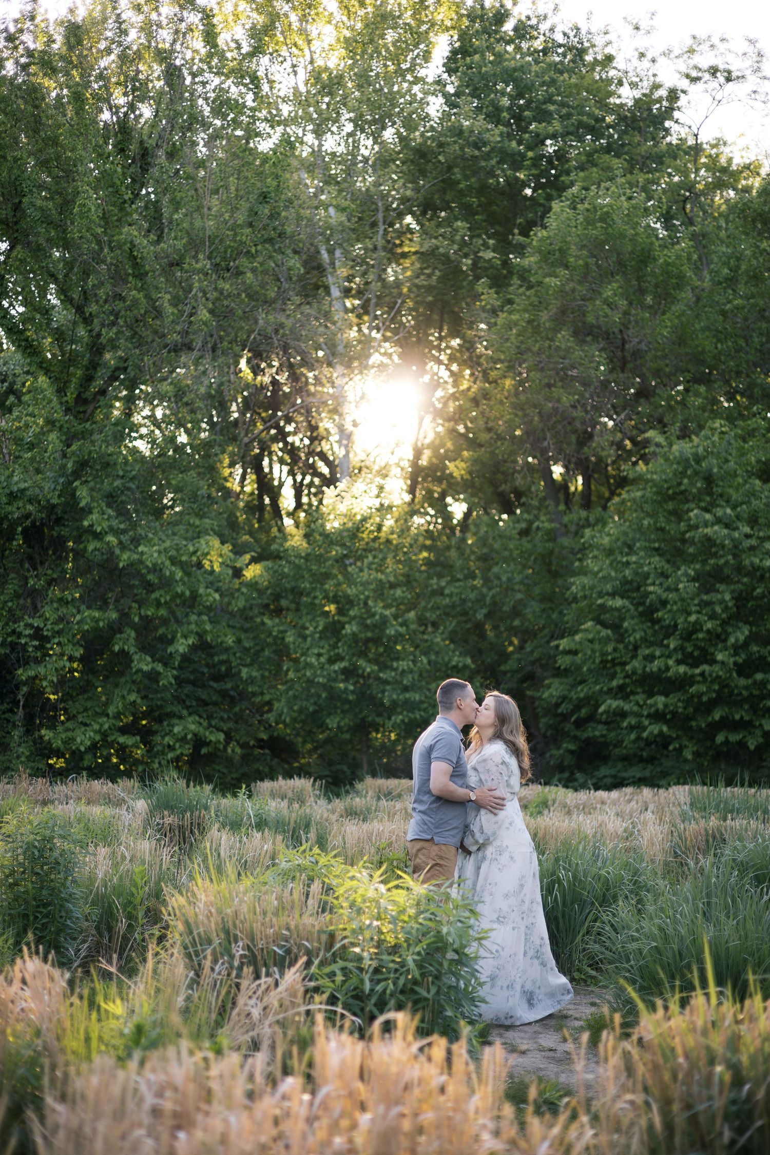 Couple sharing a romantic kiss in a sunlit field surrounded by lush green trees at maternity session by Katy Rose Photography.