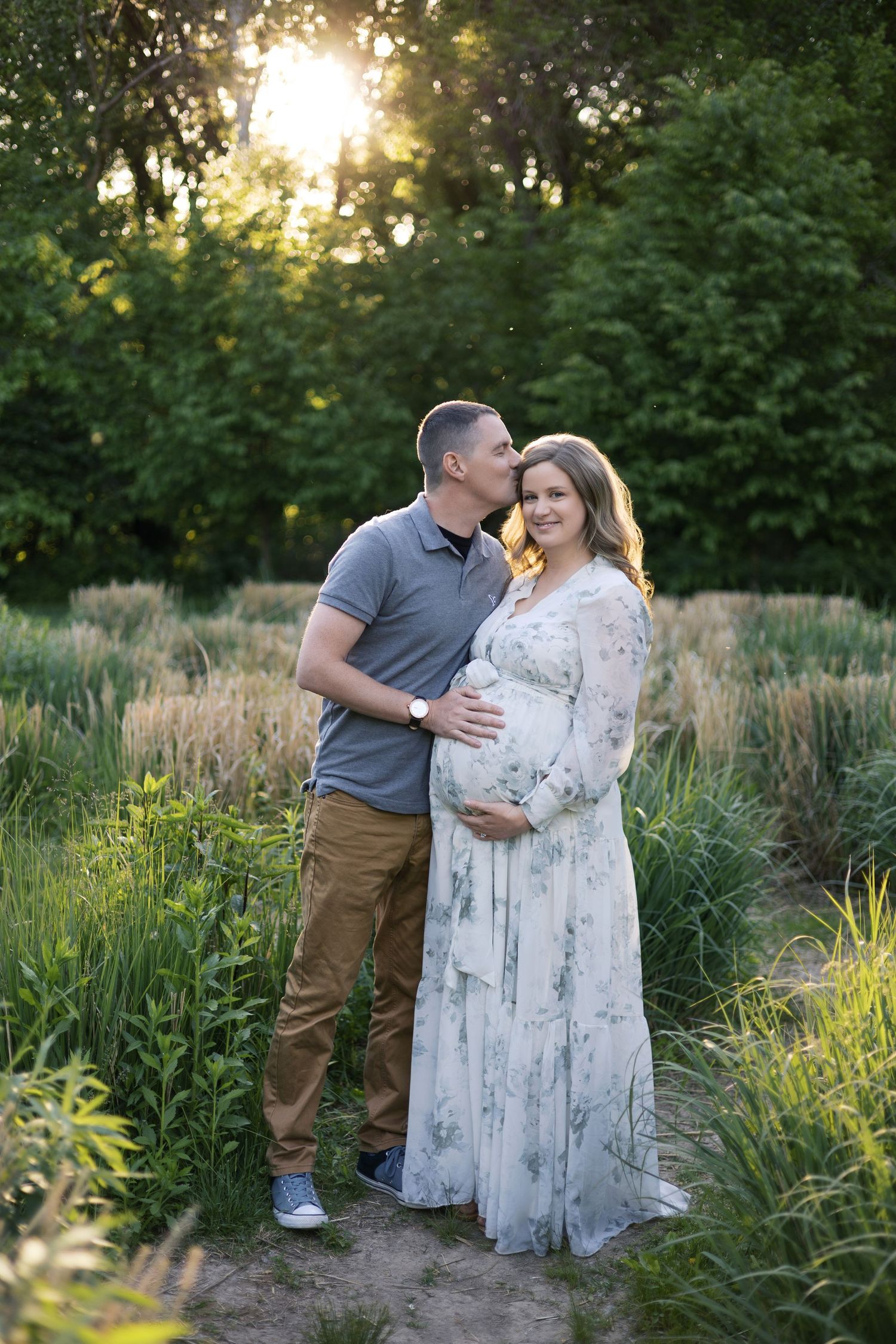 Man kissing pregnant woman's cheek while embracing her baby bump in sunlit meadow at sunset by Katy Rose Photography.