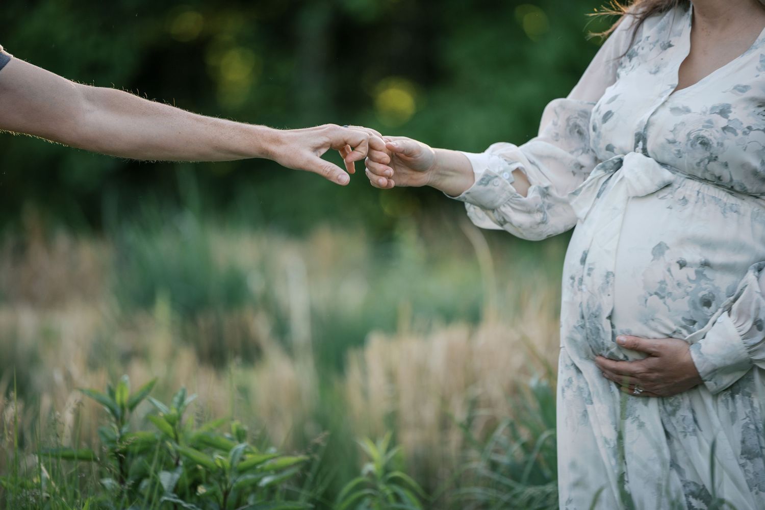 Pregnant woman in floral dress holds hands with partner outdoors in lush green field during maternity photo session by Katy Rose Photography