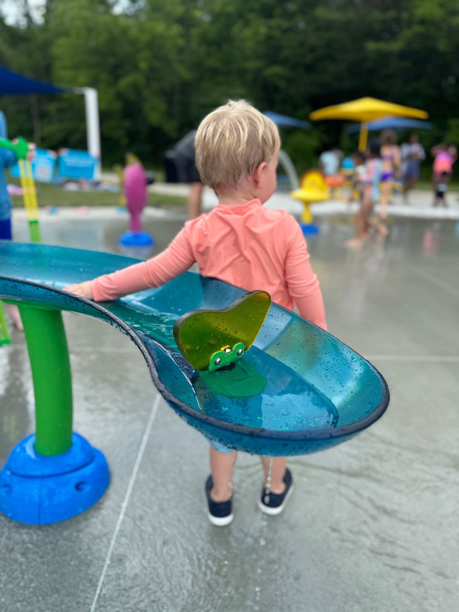 Young blonde child in pink rash guard playing at colorful outdoor splash pad water park on summer day.
