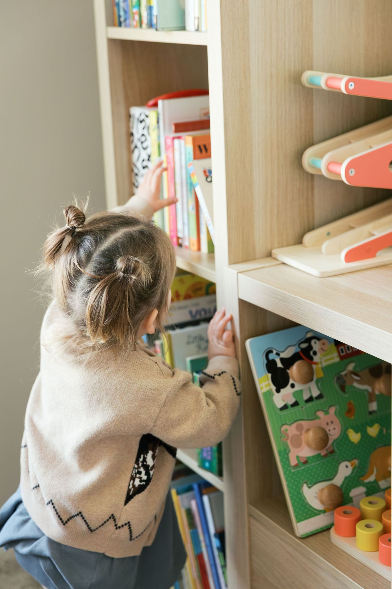 Young toddler girl reaching for books on a wooden bookshelf filled with colorful children's books and puzzles.