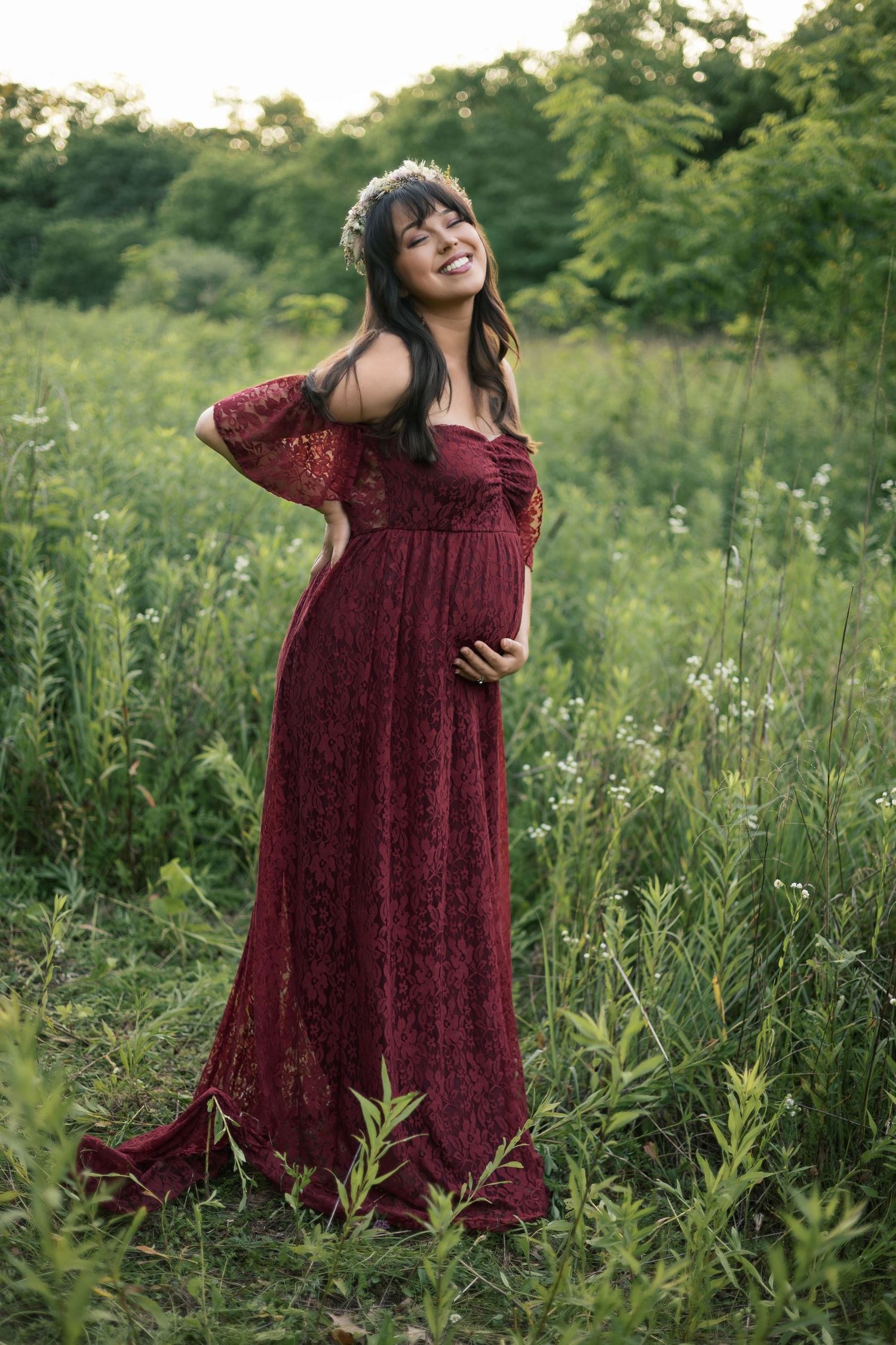 Smiling pregnant woman in burgundy lace gown with floral crown posing in a wildflower meadow.