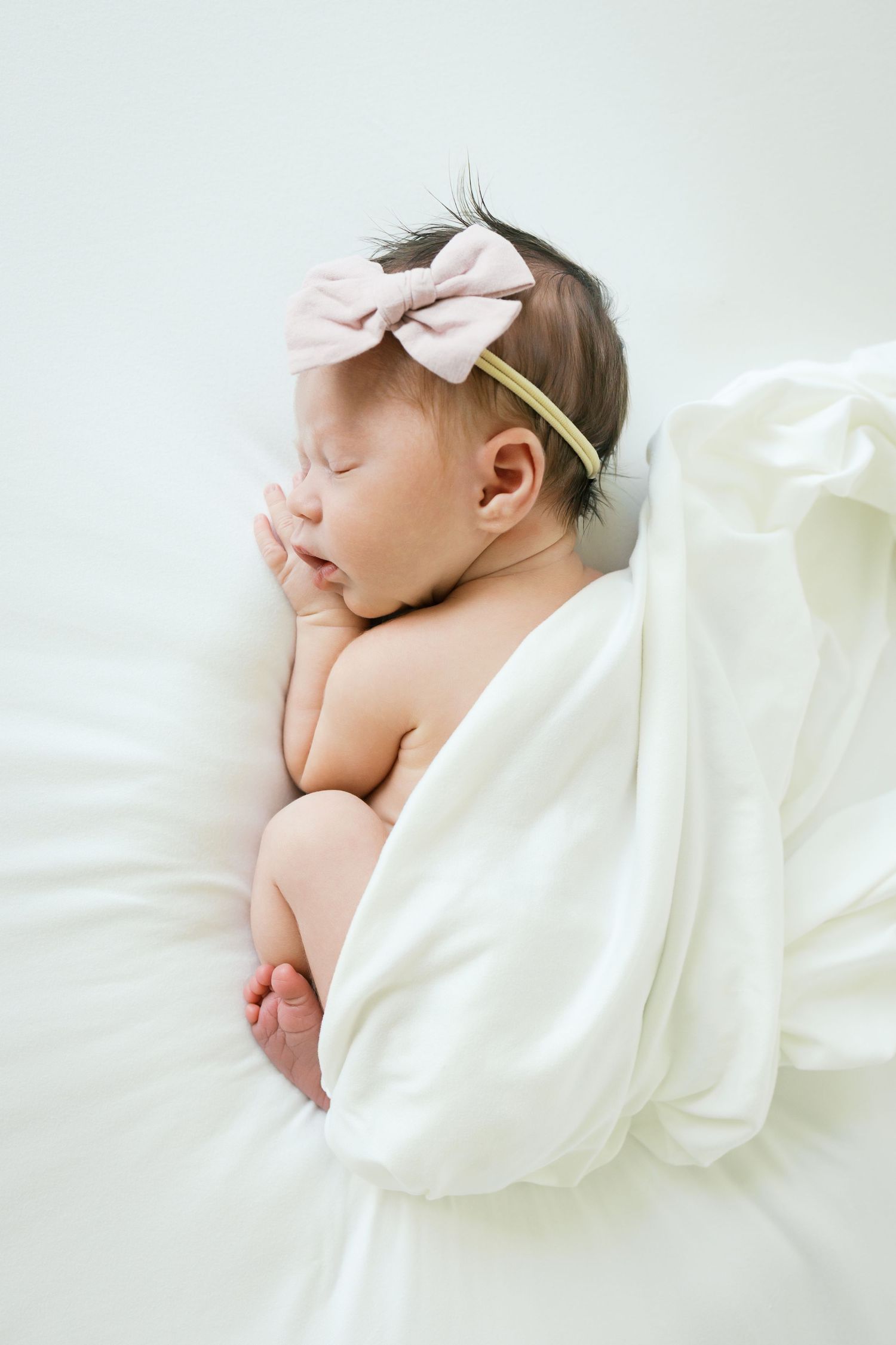 Peaceful newborn baby girl sleeping wrapped in white blanket wearing a pink bow headband.