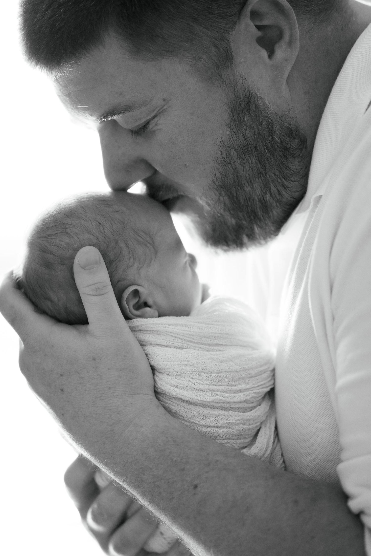 Black and white photo of a father tenderly kissing his sleeping newborn baby during in-home newborn photoshoot in Brownsburg, Indiana
