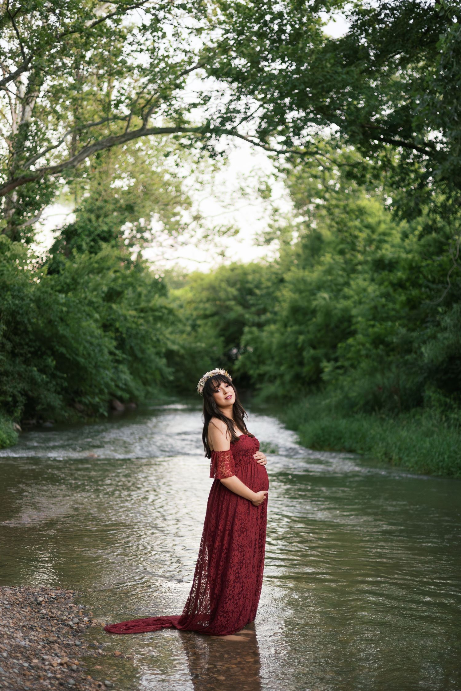 Pregnant woman in red lace off-shoulder gown standing in a shallow creek surrounded by lush green trees.