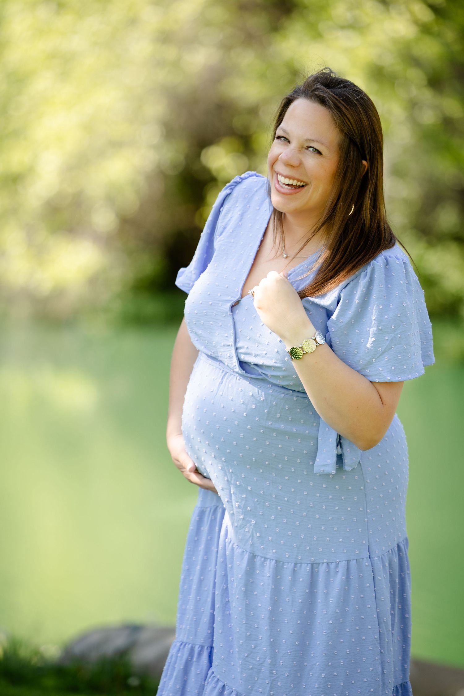 Smiling pregnant woman in blue dress, cradling her baby bump for maternity session in Avon, Indiana at Avon Gardens by Katy Rose Photography