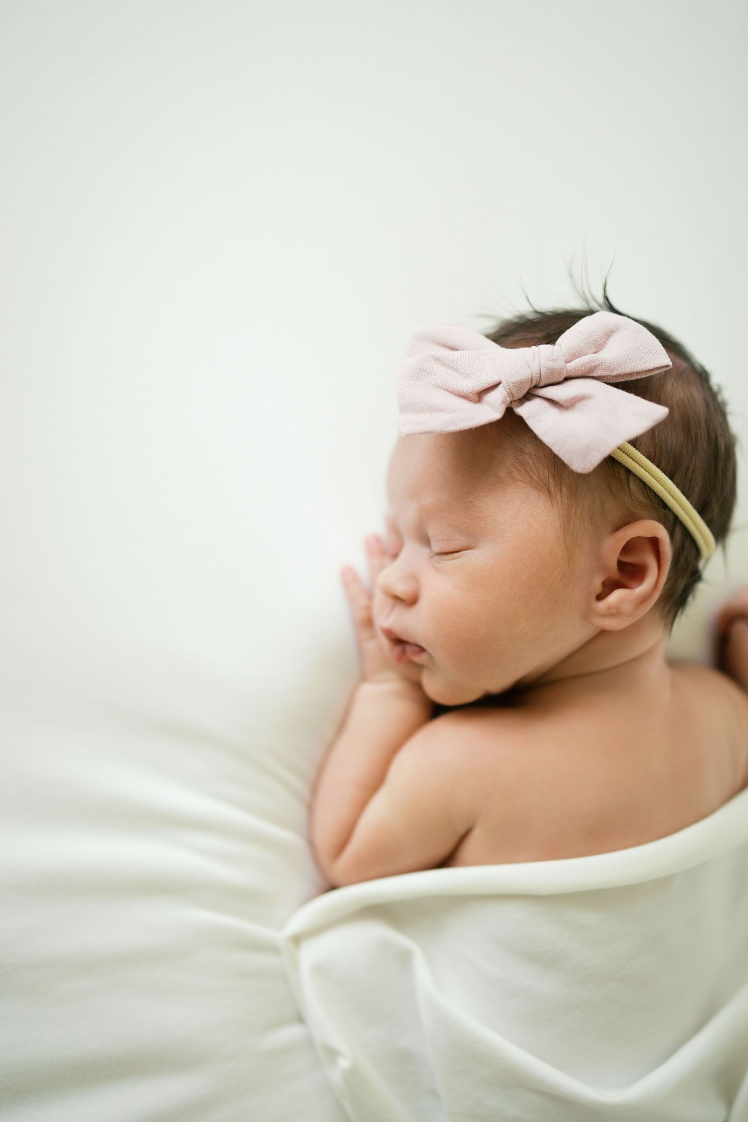 Sleeping newborn baby girl with pink bow headband wrapped in white blanket.