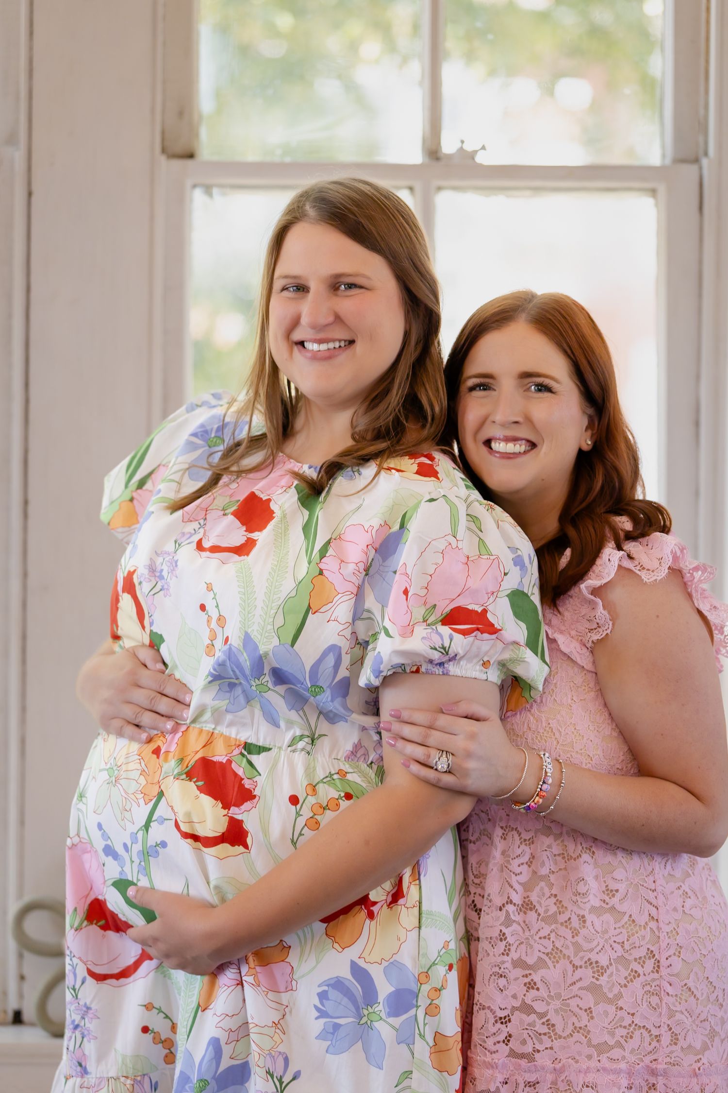 Two expecting mothers share joyful moment by window in floral and pink dresses.